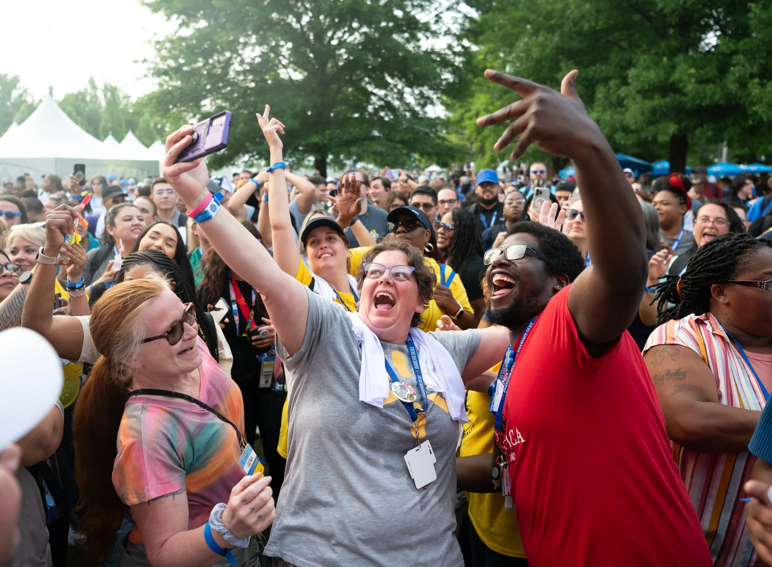 A diverse group of people celebrating outdoors at an event, smiling, with some raising their hands and taking photos, surrounded by trees and tents.