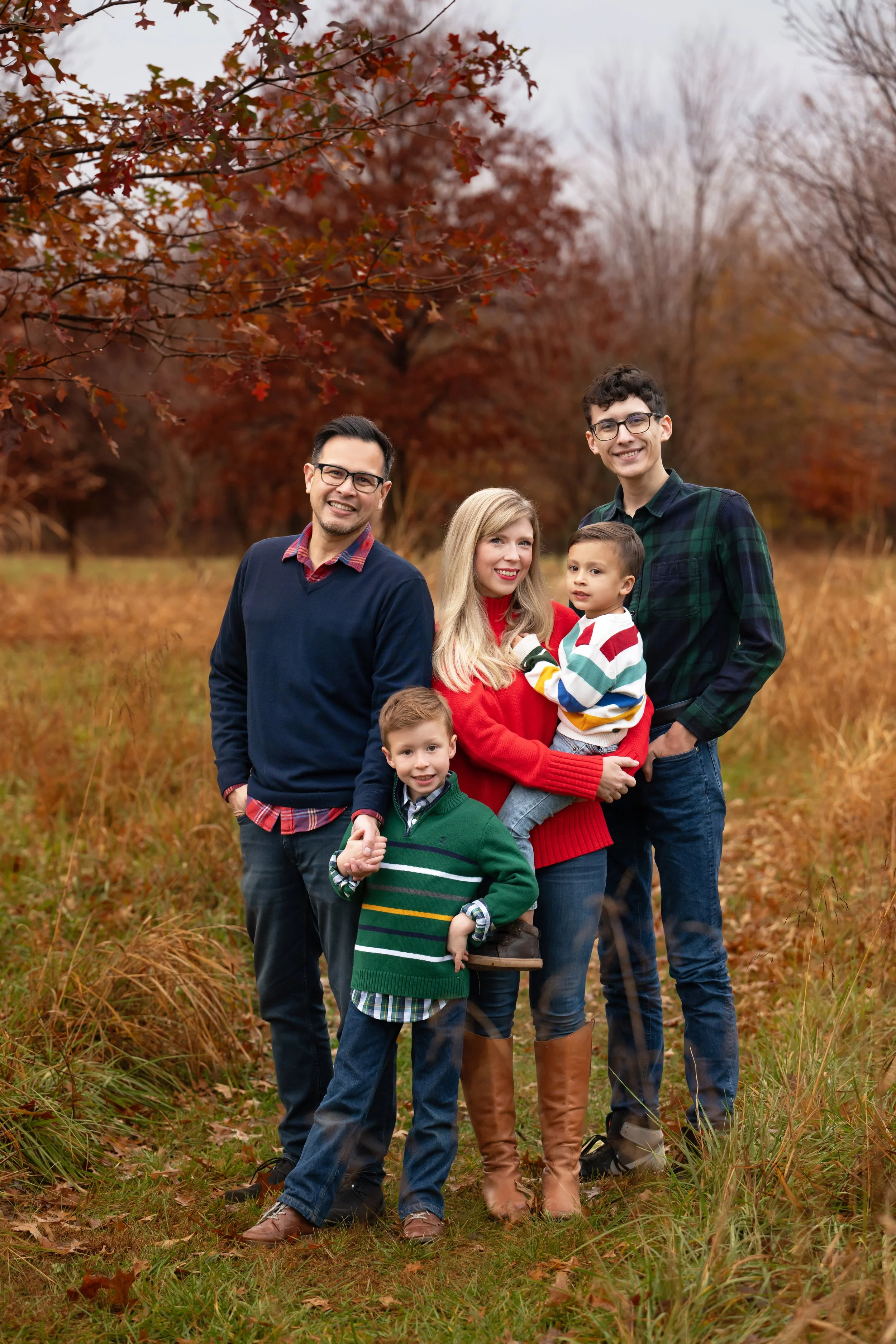 A family of five standing in a park with fall foliage, smiling at the camera. The family includes two men, one woman, and two boys, all dressed in warm clothing.