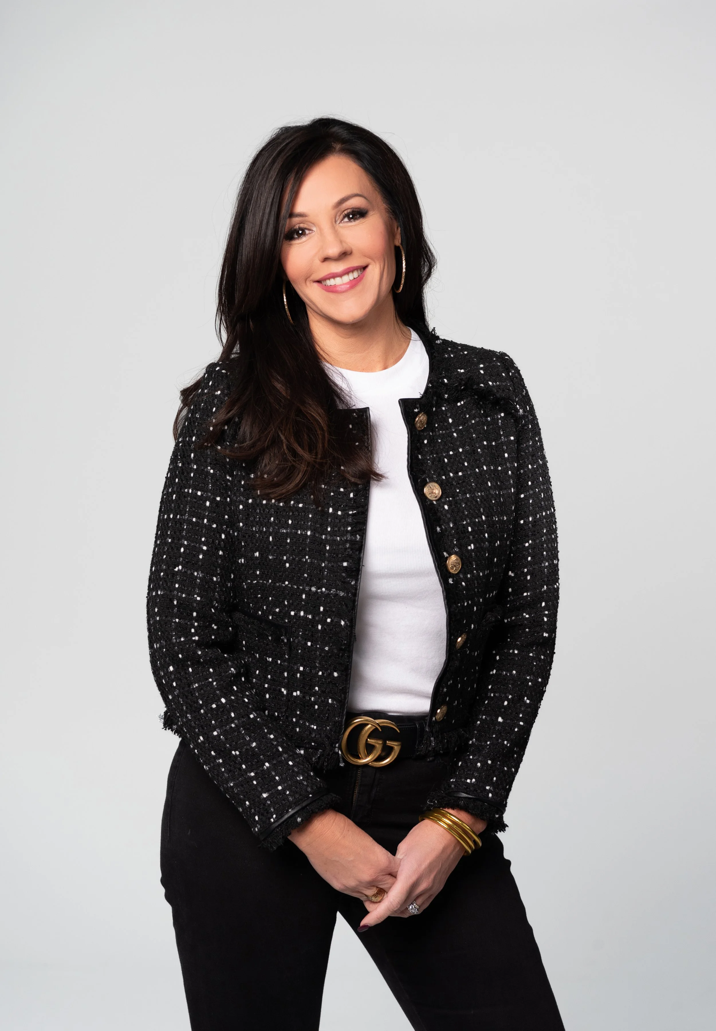 A woman with long dark hair, wearing gold hoop earrings, a black and white Tweed jacket, white top, black pants, and a "GG" belt, standing against a plain white background, smiling and looking at the camera.