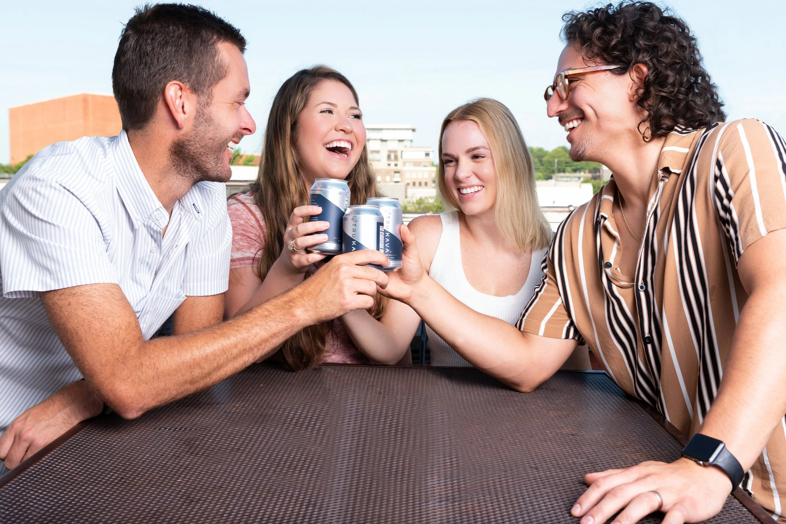 Group of five friends sitting outside on a rooftop, smiling, holding and clinking cans of beverages, with buildings and blue sky in the background.
