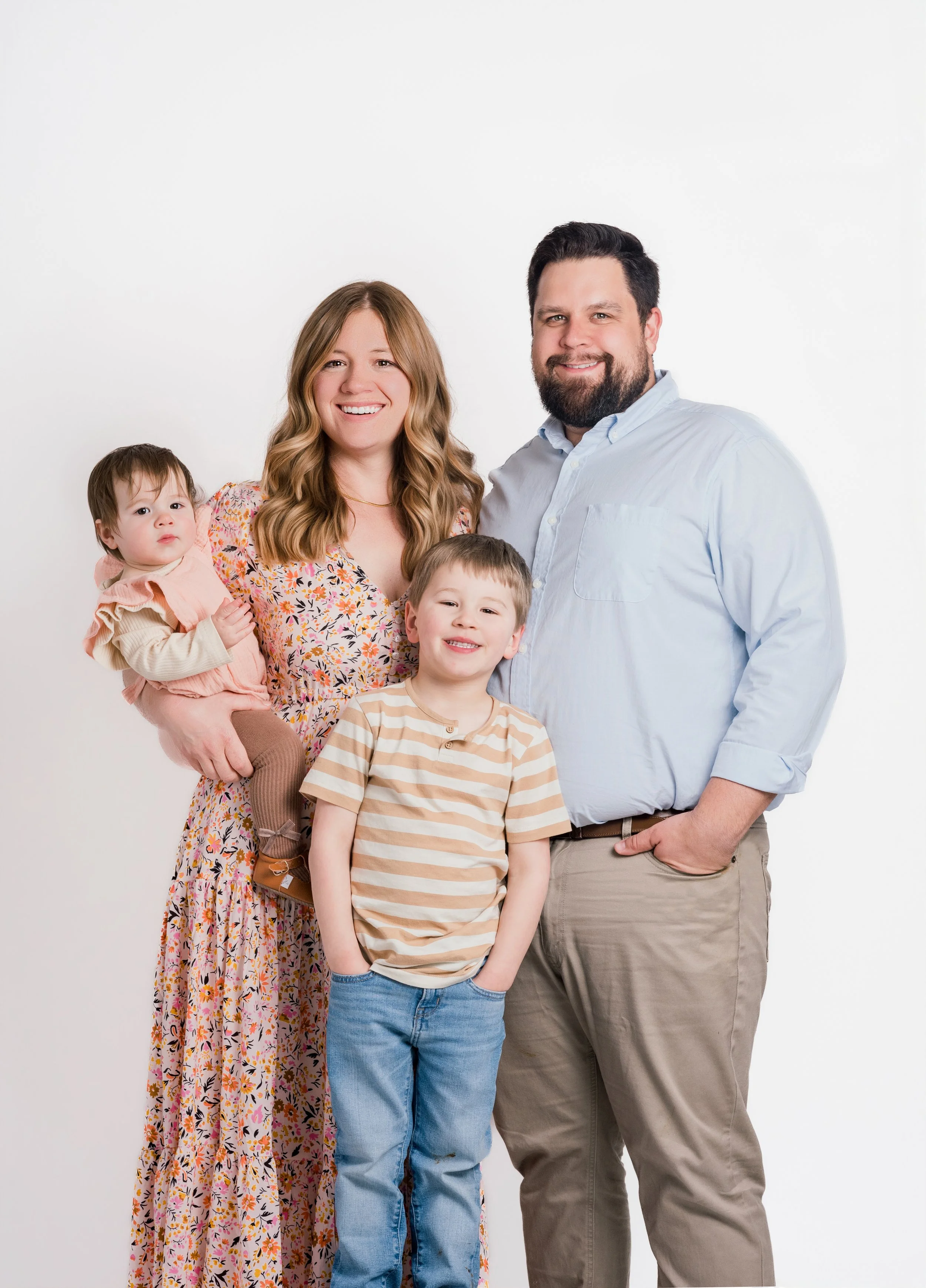 Family portrait of four, including a woman with wavy blonde hair, a man with dark hair and beard, a young boy with short hair, and a young girl with dark hair, posing against a white background.