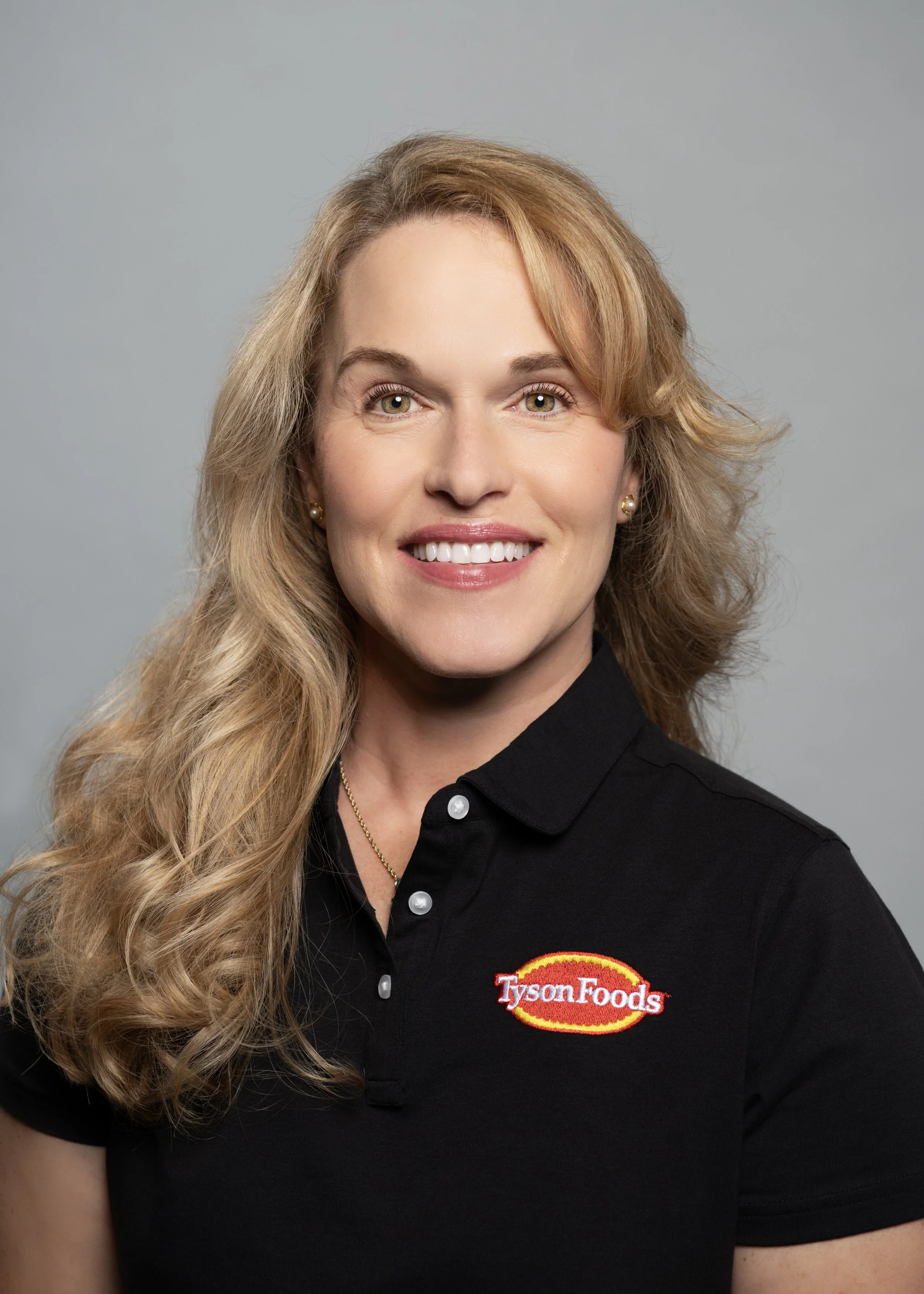 A smiling woman with wavy blonde hair wearing a black polo shirt with a Tyson Foods logo on it, against a gray background.