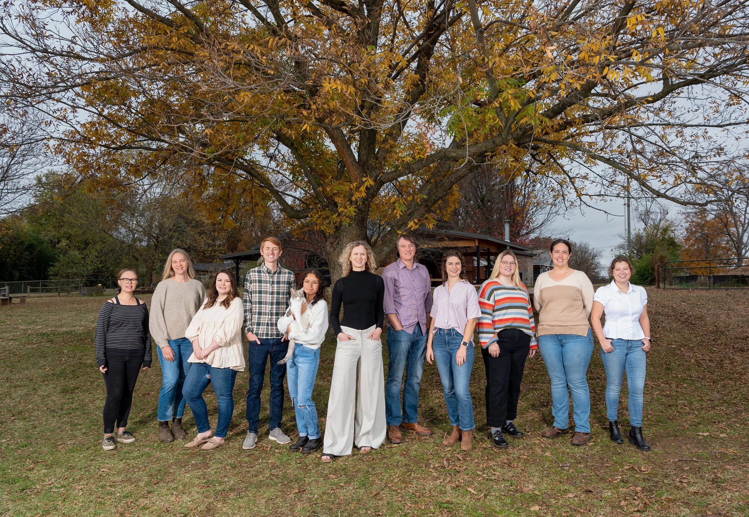 Group of twelve diverse people standing outside in front of a large tree with autumn leaves, smiling at the camera.