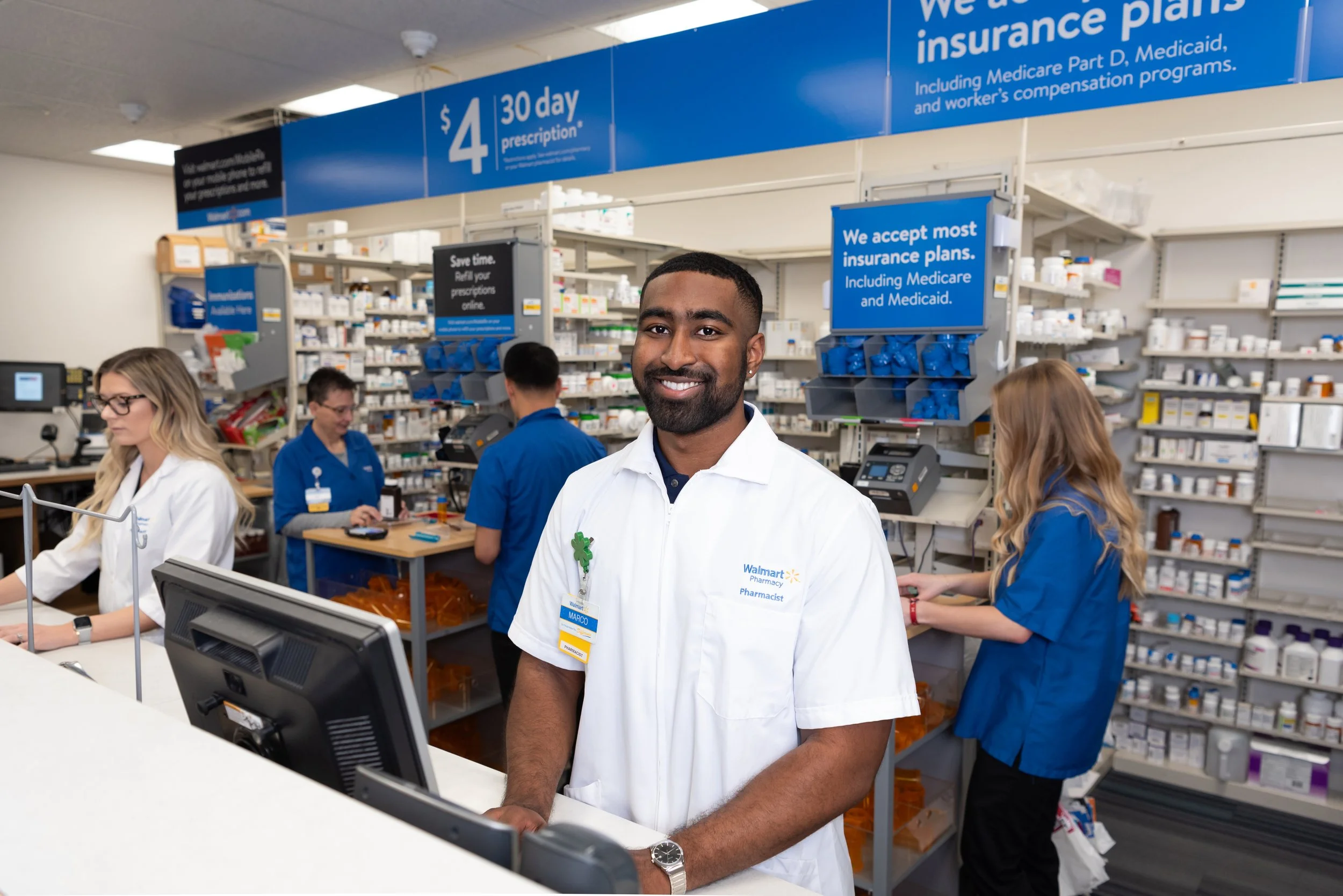 A smiling male pharmacist standing at a pharmacy counter with shelves of medications and pharmacy staff in the background.