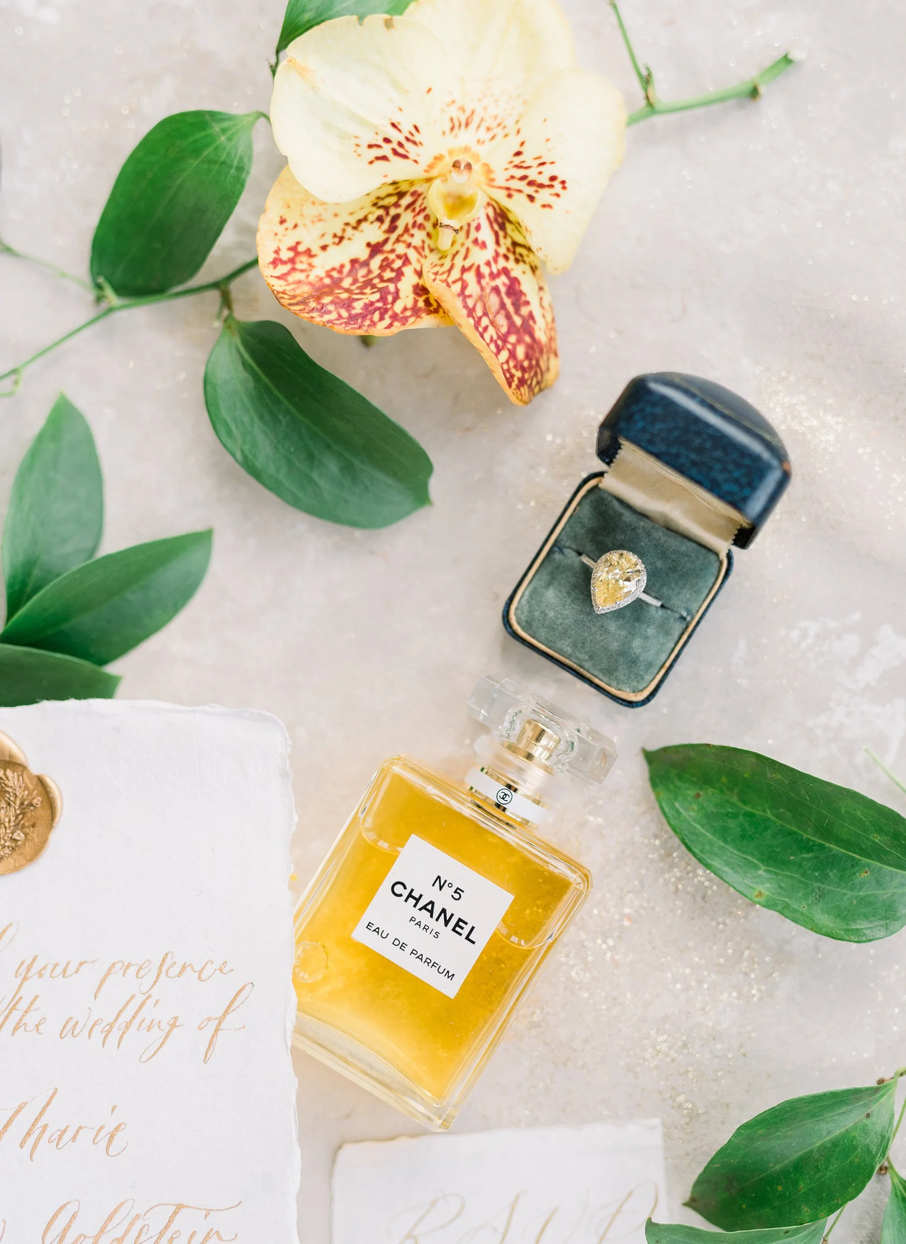 A yellow Chanel No. 5 perfume bottle, a jewelry box with a diamond ring, a wedding invitation, a white card, green leaves, and a white orchid flower on a light-colored surface.