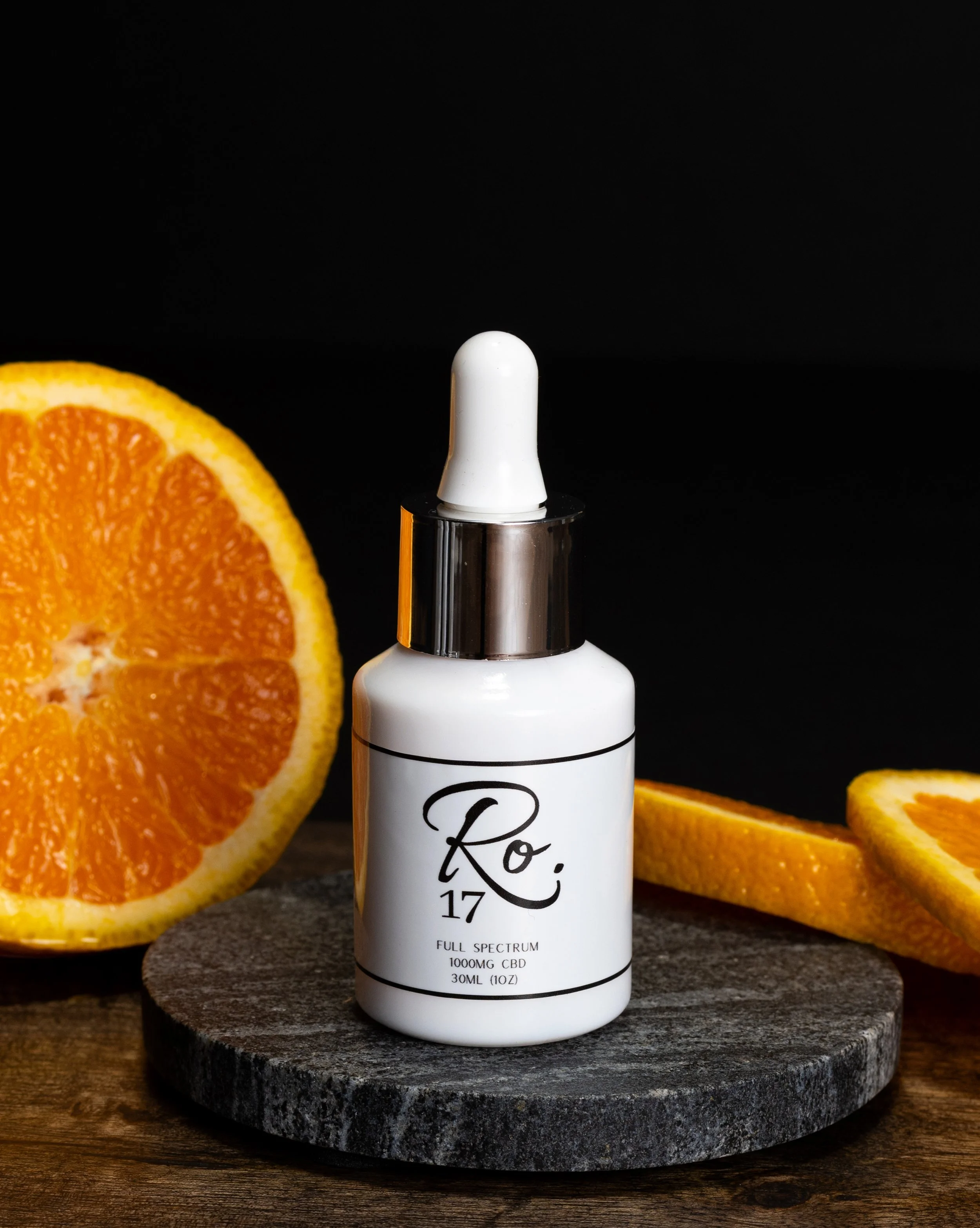 A white bottle of CBD serum with a dropper cap sits on a round gray stone platform surrounded by slices of orange, against a black background.