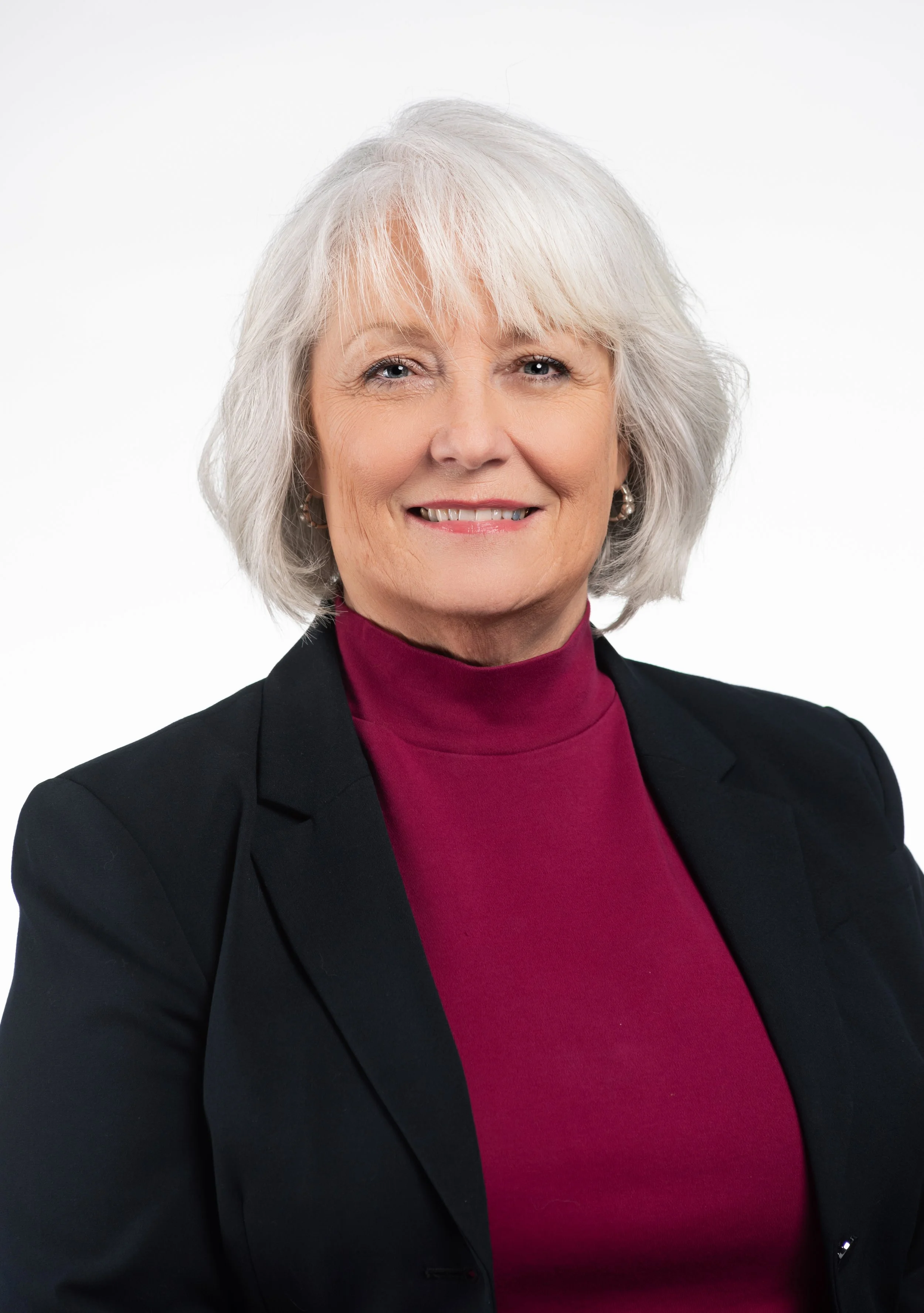 A portrait of an older woman with gray hair, wearing a black blazer and a magenta turtleneck, smiling against a white background.