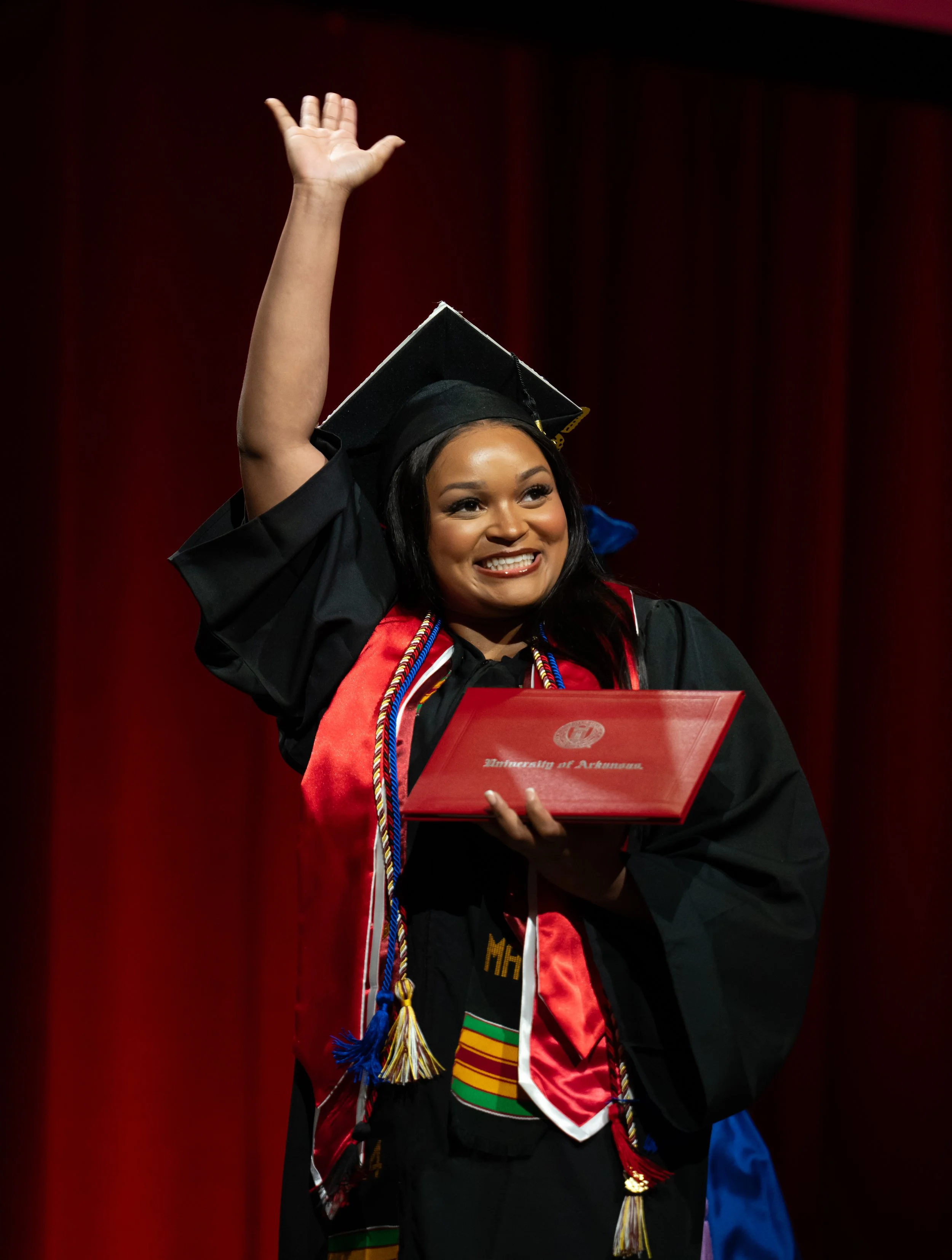 A woman in a graduation cap and gown holding a diploma and waving on stage. Red curtain background.