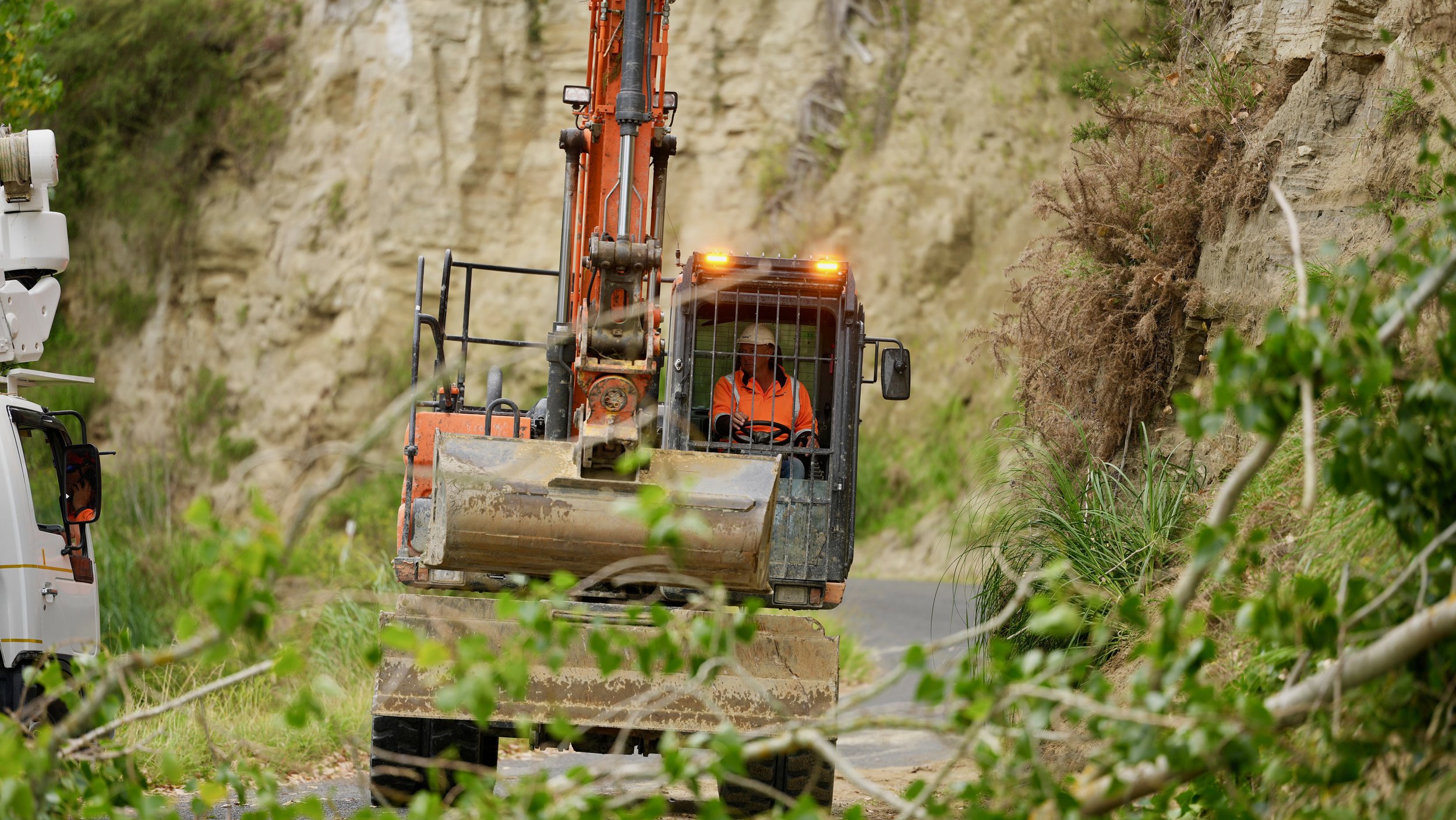 A construction worker driving an orange excavator on a dirt road beside a rocky hillside, surrounded by greenery.