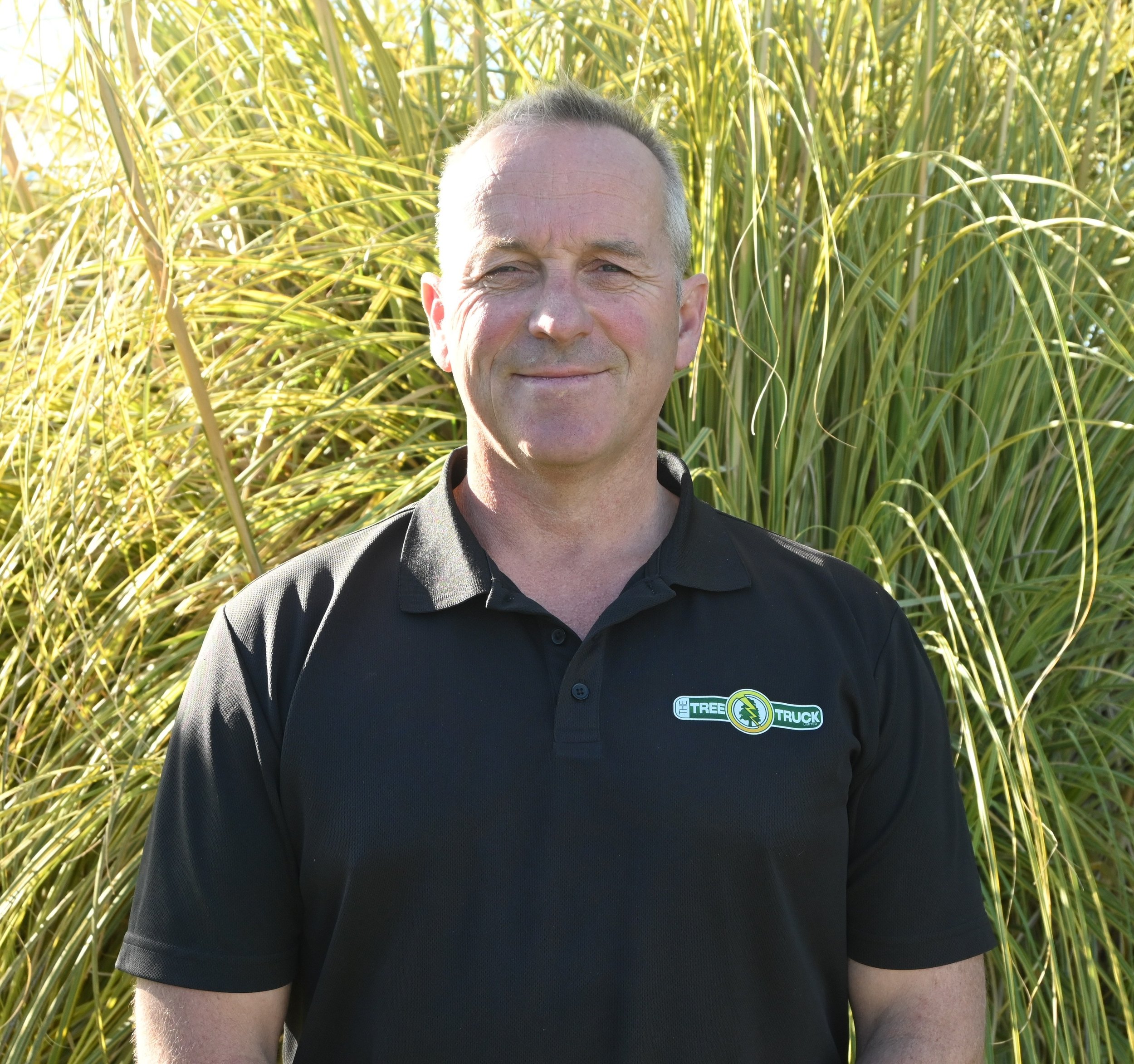 A man wearing a black polo shirt with a Tree Truck logo standing outdoors in front of green plants