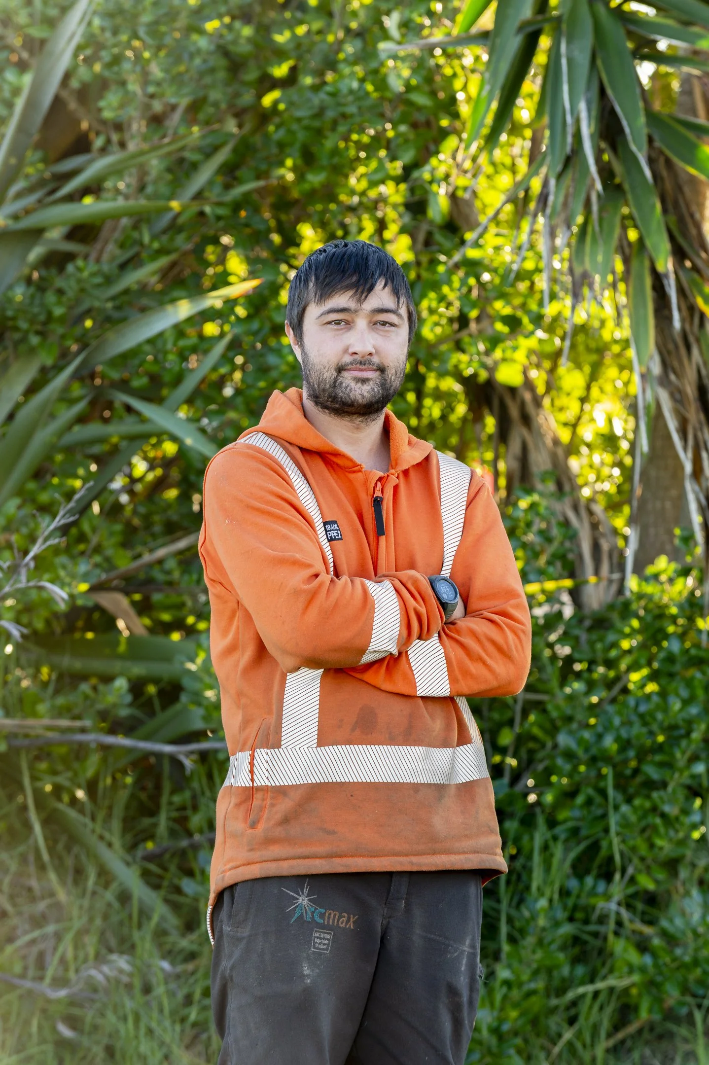 A man with dark hair and a beard, smiling, wearing an orange high-visibility work shirt, standing outdoors in front of tall, yellowish-green decorative grasses.