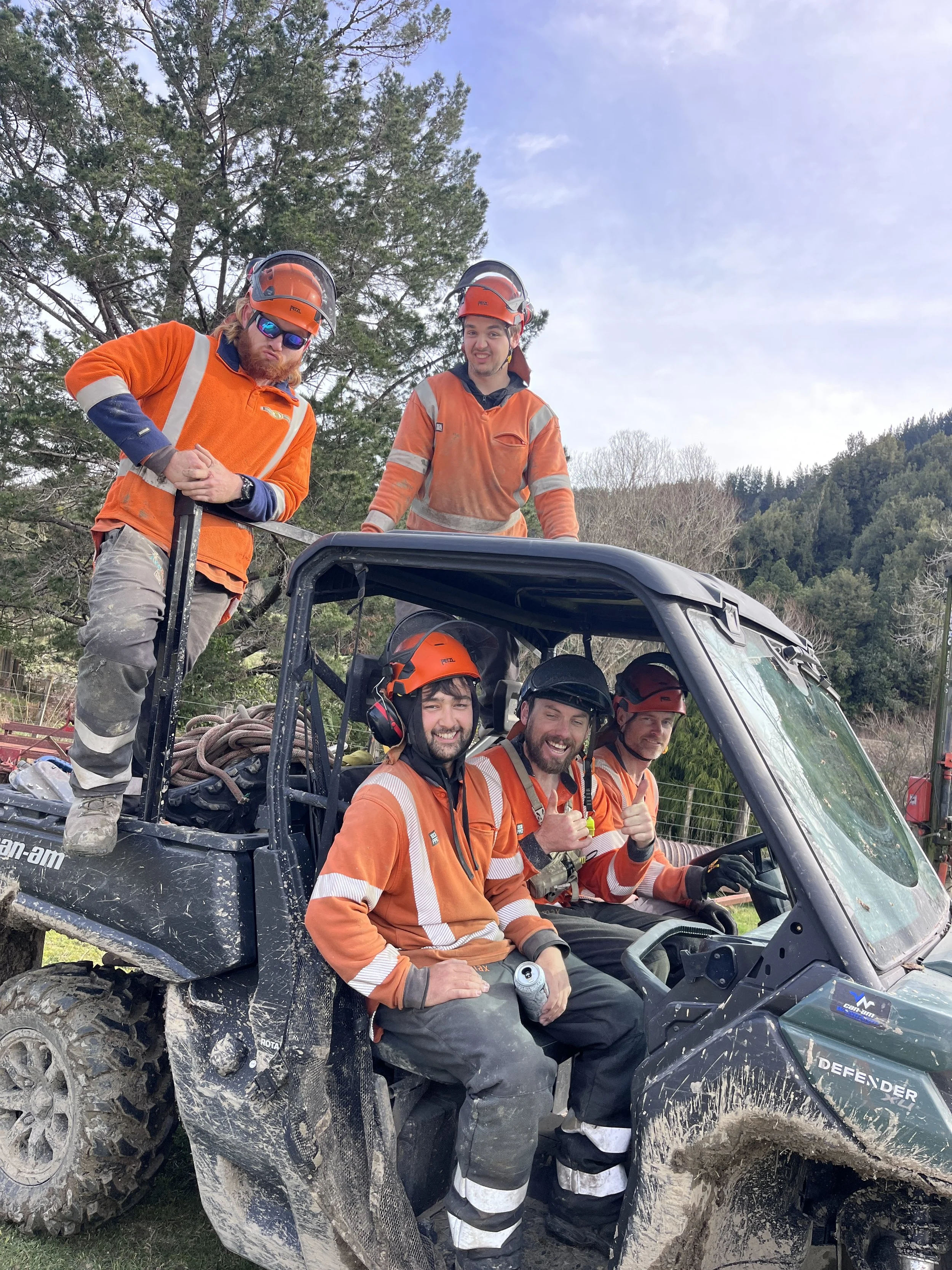 Group of five workers in orange safety uniforms, helmets, and protective gear sitting and standing on a muddy all-terrain vehicle outdoors with trees and hills in the background.