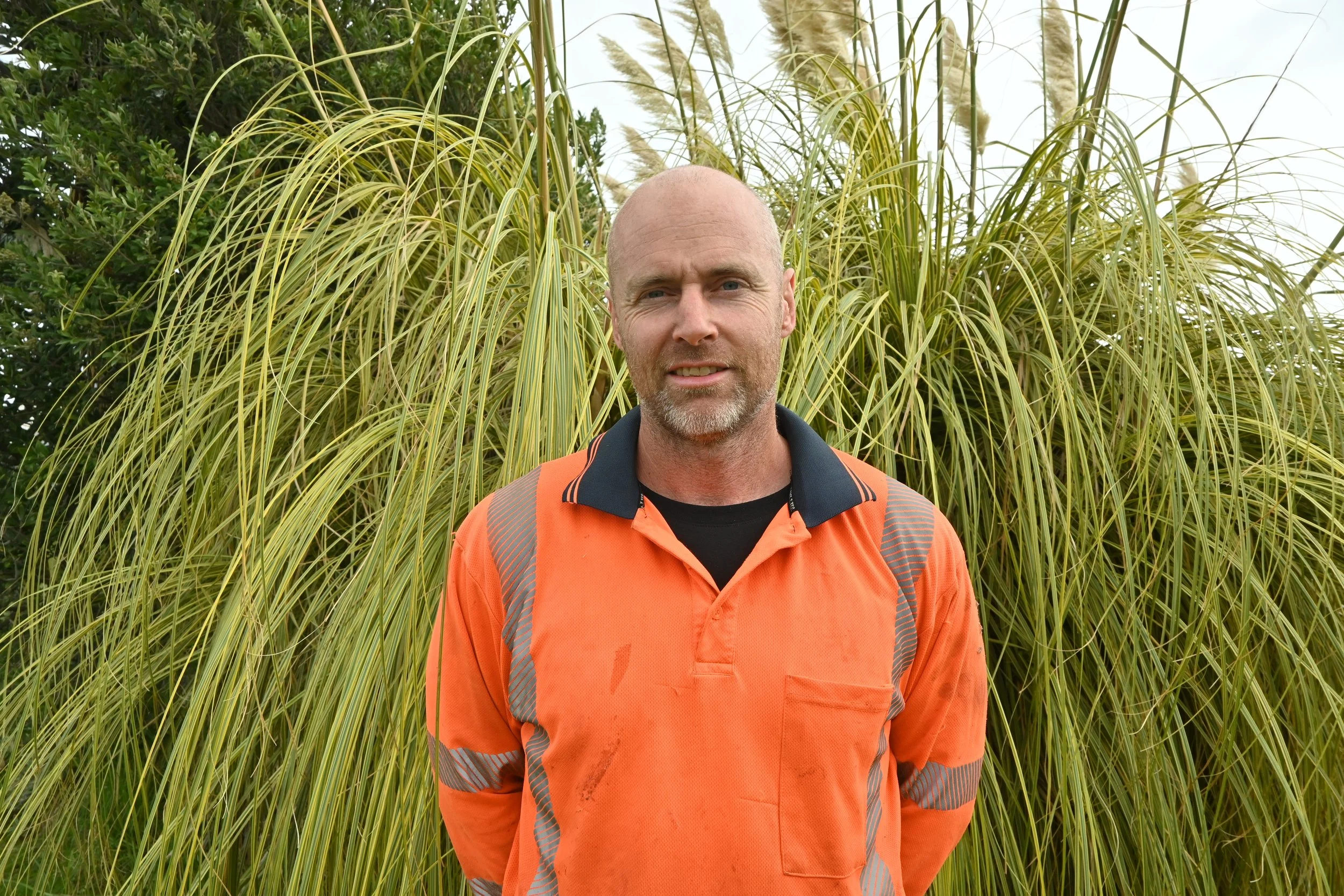 A man with a beard and short hair wearing an orange work shirt standing in front of tall green grass.
