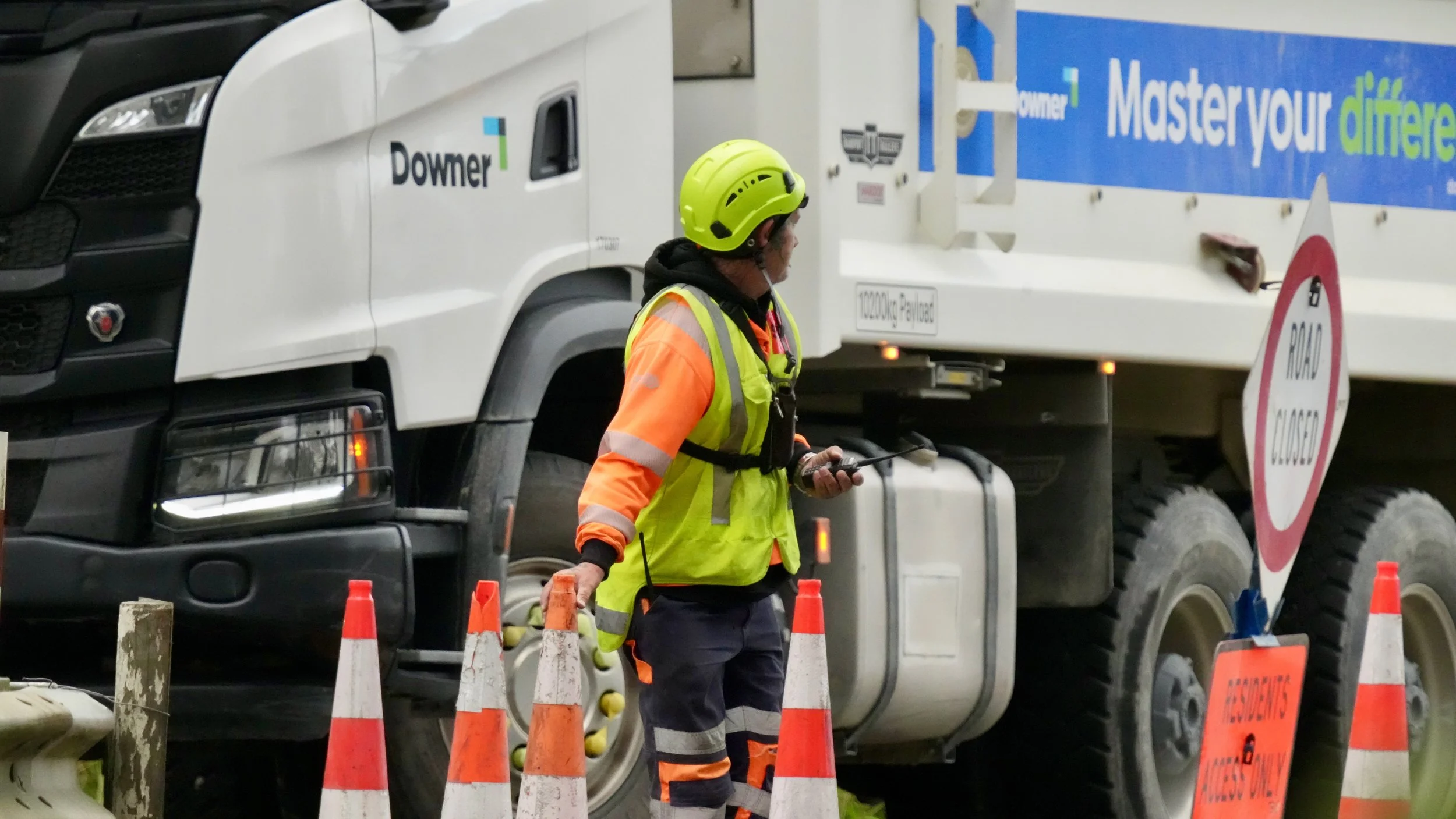 A construction worker in safety gear, including a yellow helmet and reflective orange and yellow jacket, standing near a large white delivery truck and orange traffic cones at a road closure site.