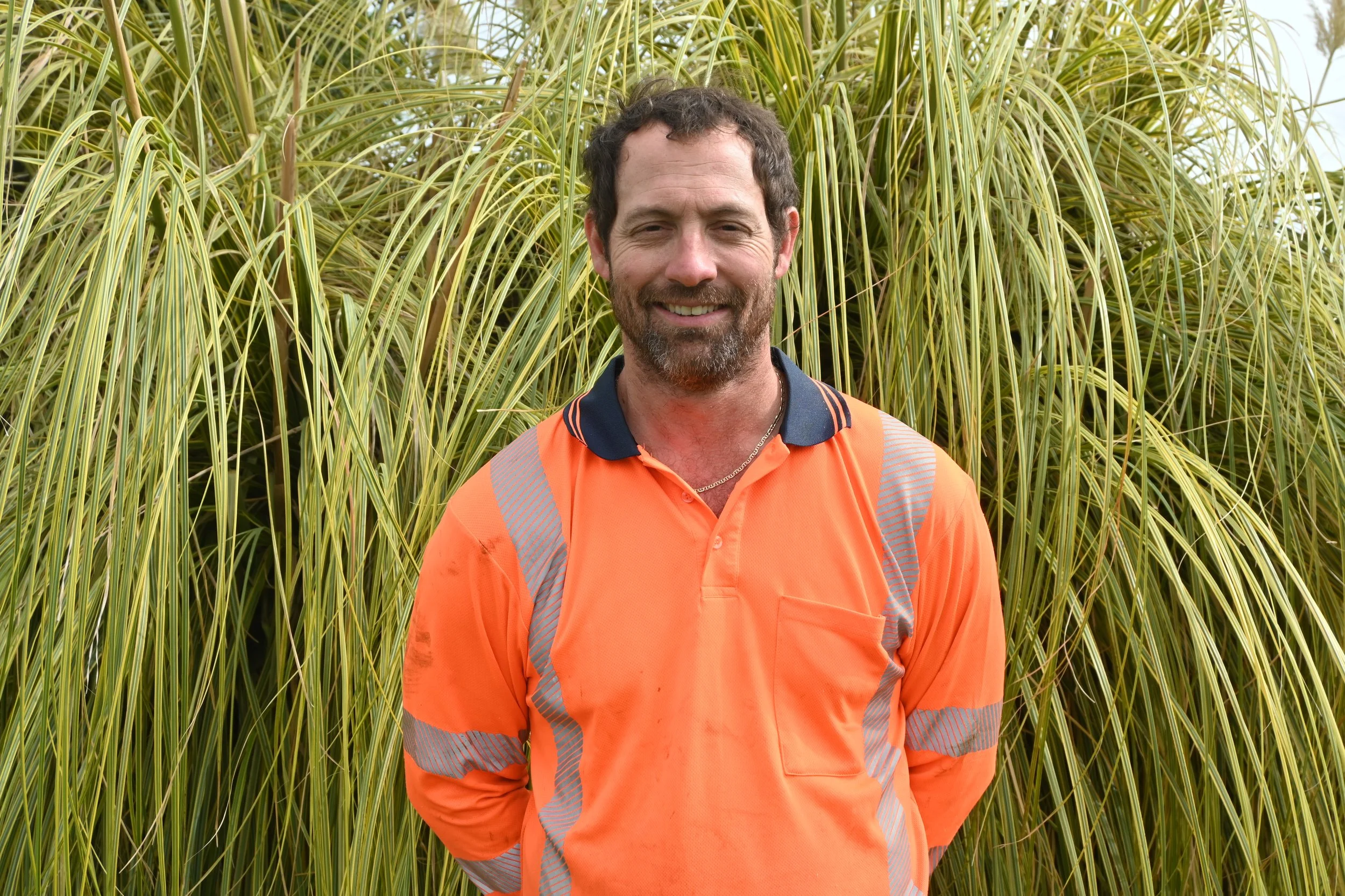 A man smiling outdoors in front of tall, green, grassy plants, wearing an orange safety shirt with reflective stripes.