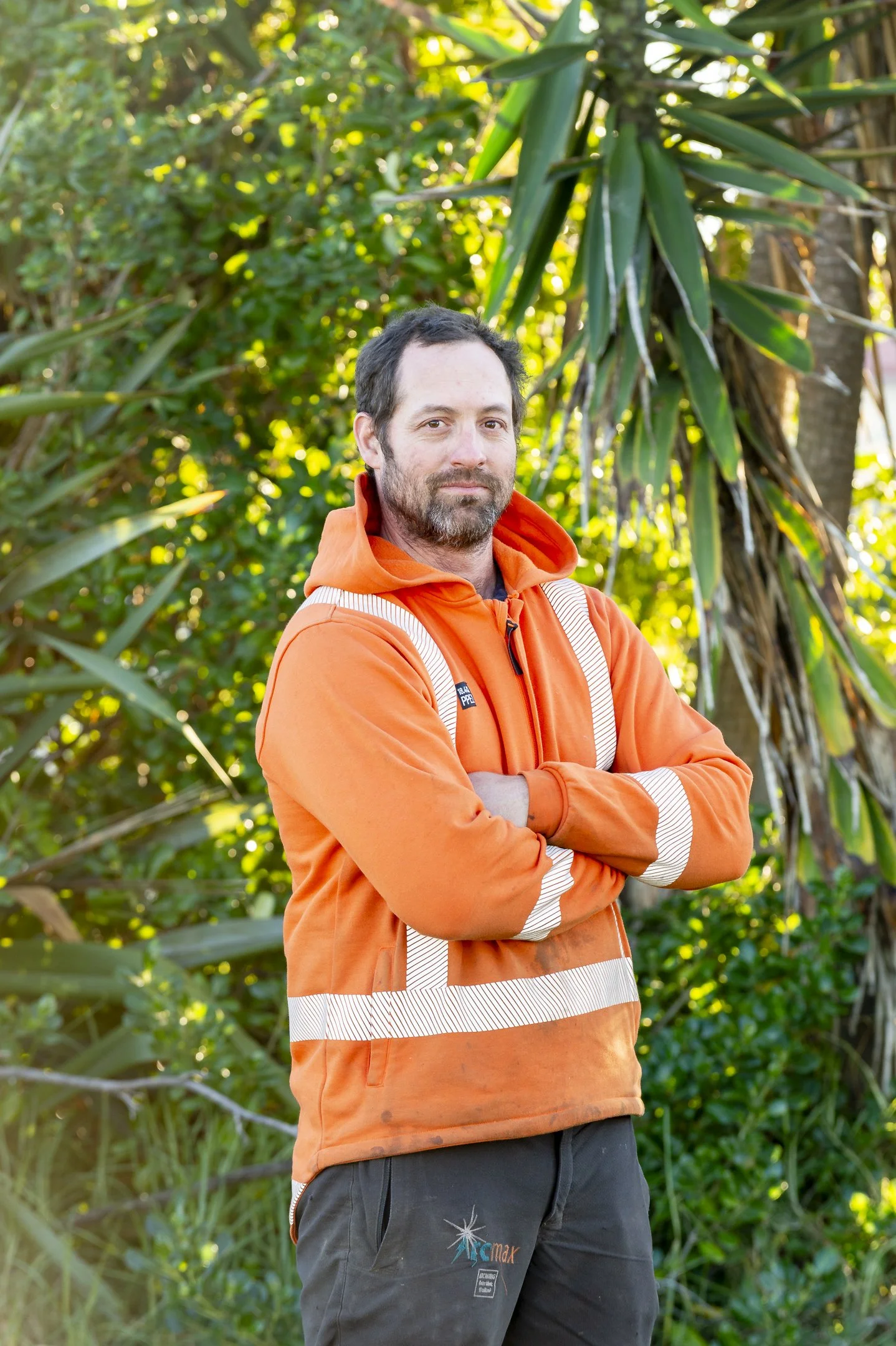 A man smiling outdoors in front of tall, green, grassy plants, wearing an orange safety shirt with reflective stripes.