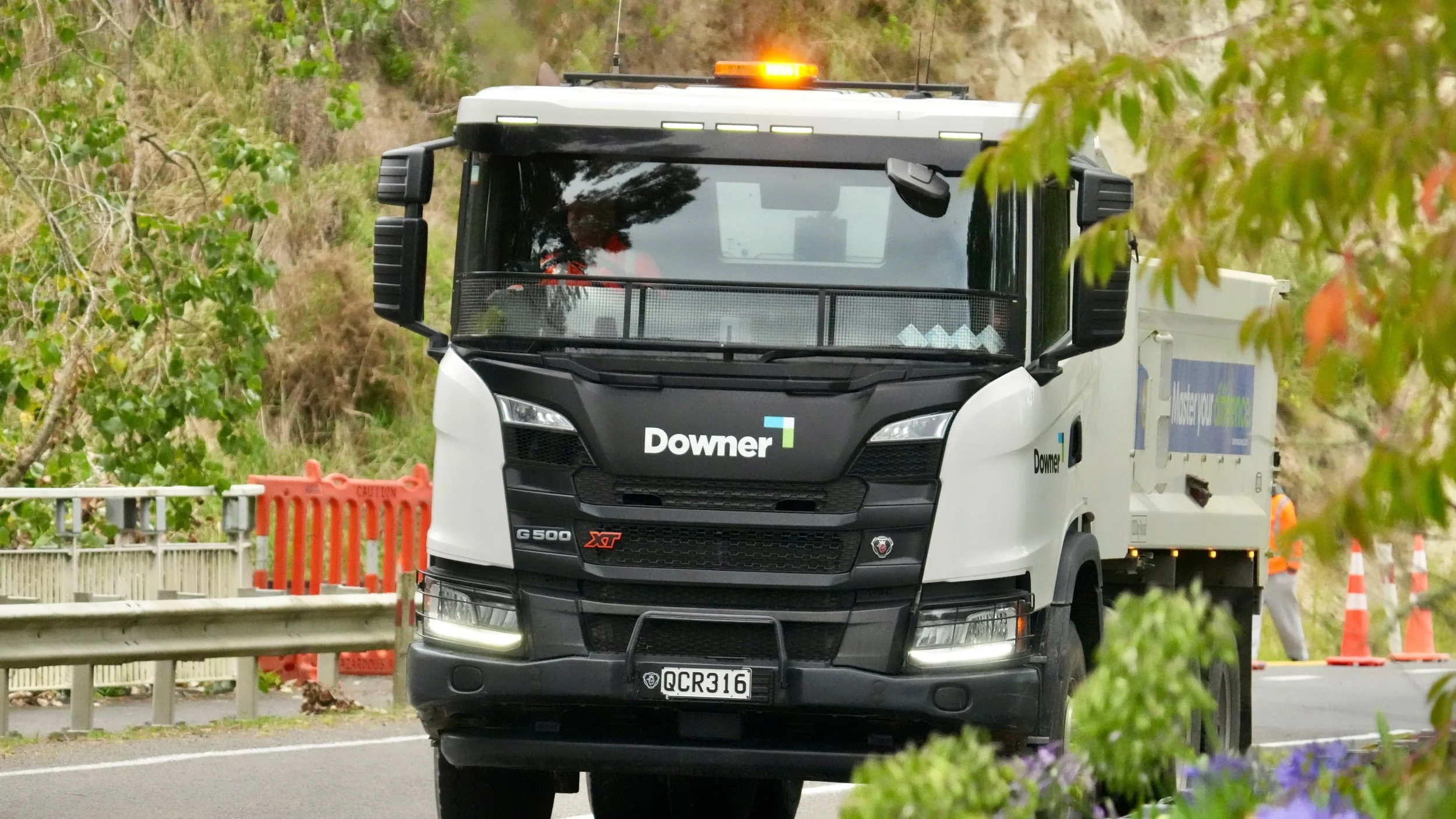 A large white utility truck with Downer branding on the front, driving on a highway with construction cones and a worker in safety gear in the background, partially obscured by green foliage and flowers.
