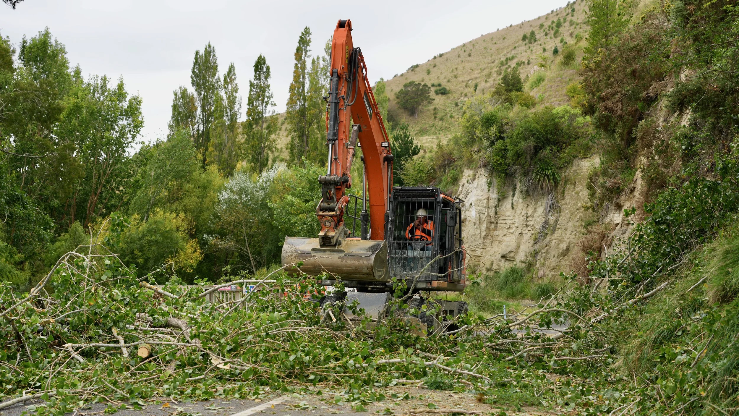 An excavator clearing fallen tree branches from a road surrounded by green trees and hills.