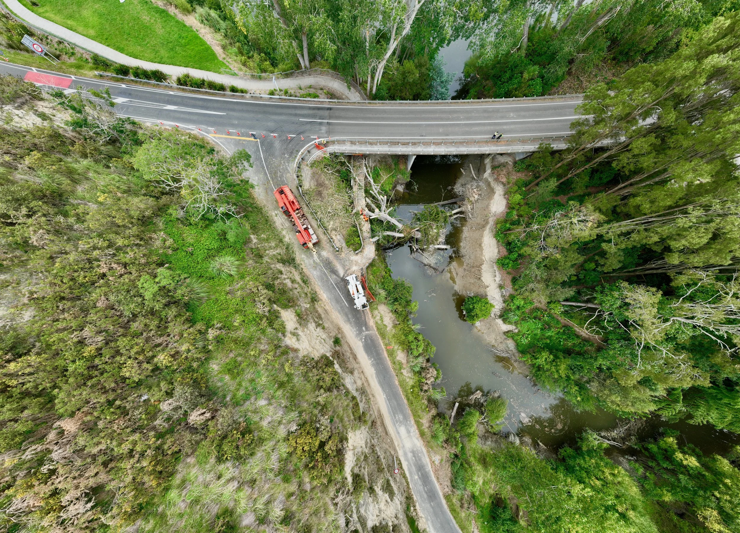 An aerial view showing emergency vehicles and personnel near a fallen tree blocking a road under a bridge over a creek in a forested area.