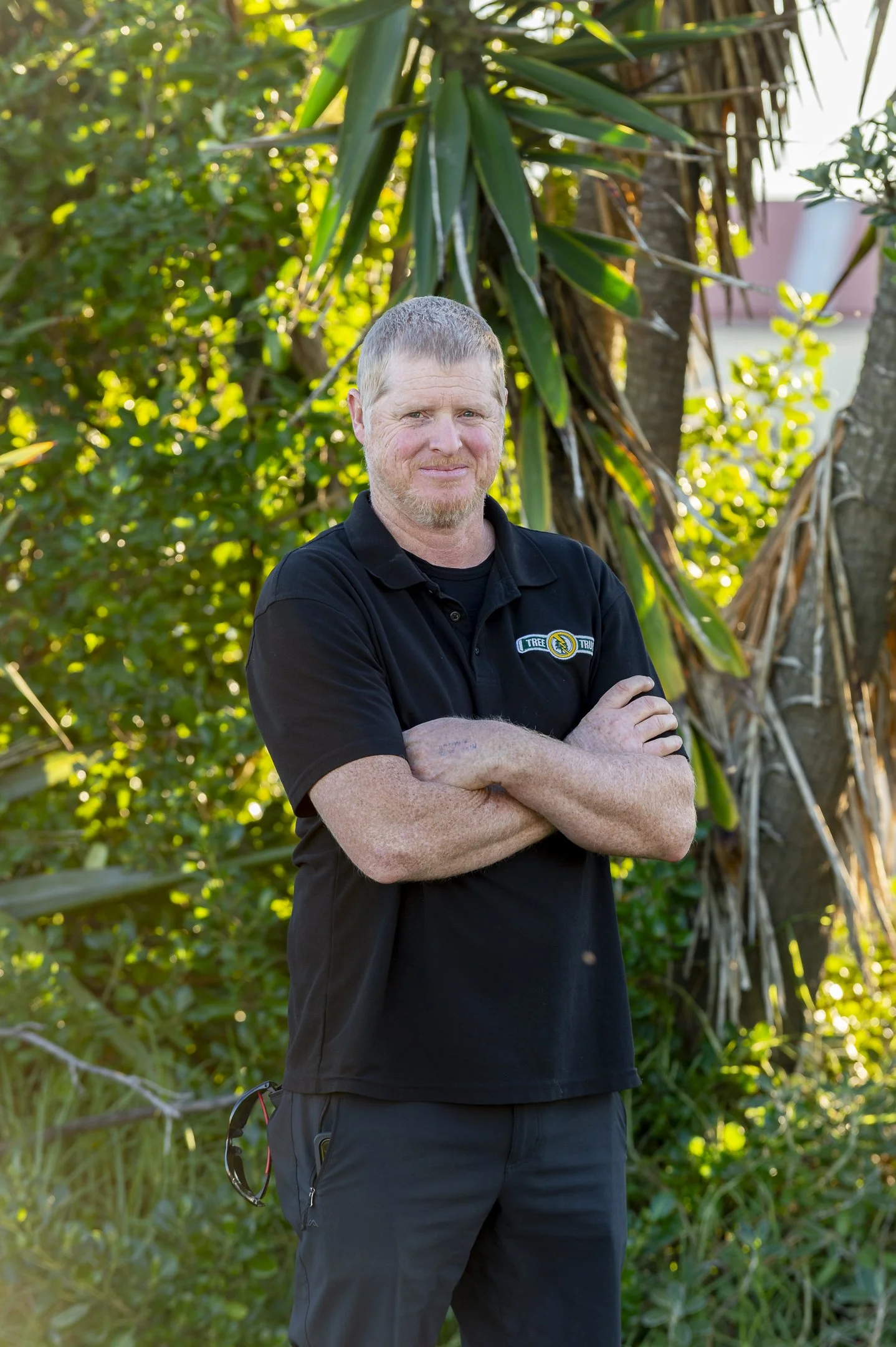 A man with short light hair and a beard standing outdoors in front of tall green plants, wearing a black polo shirt with a logo that reads 'Tree Truck.'
