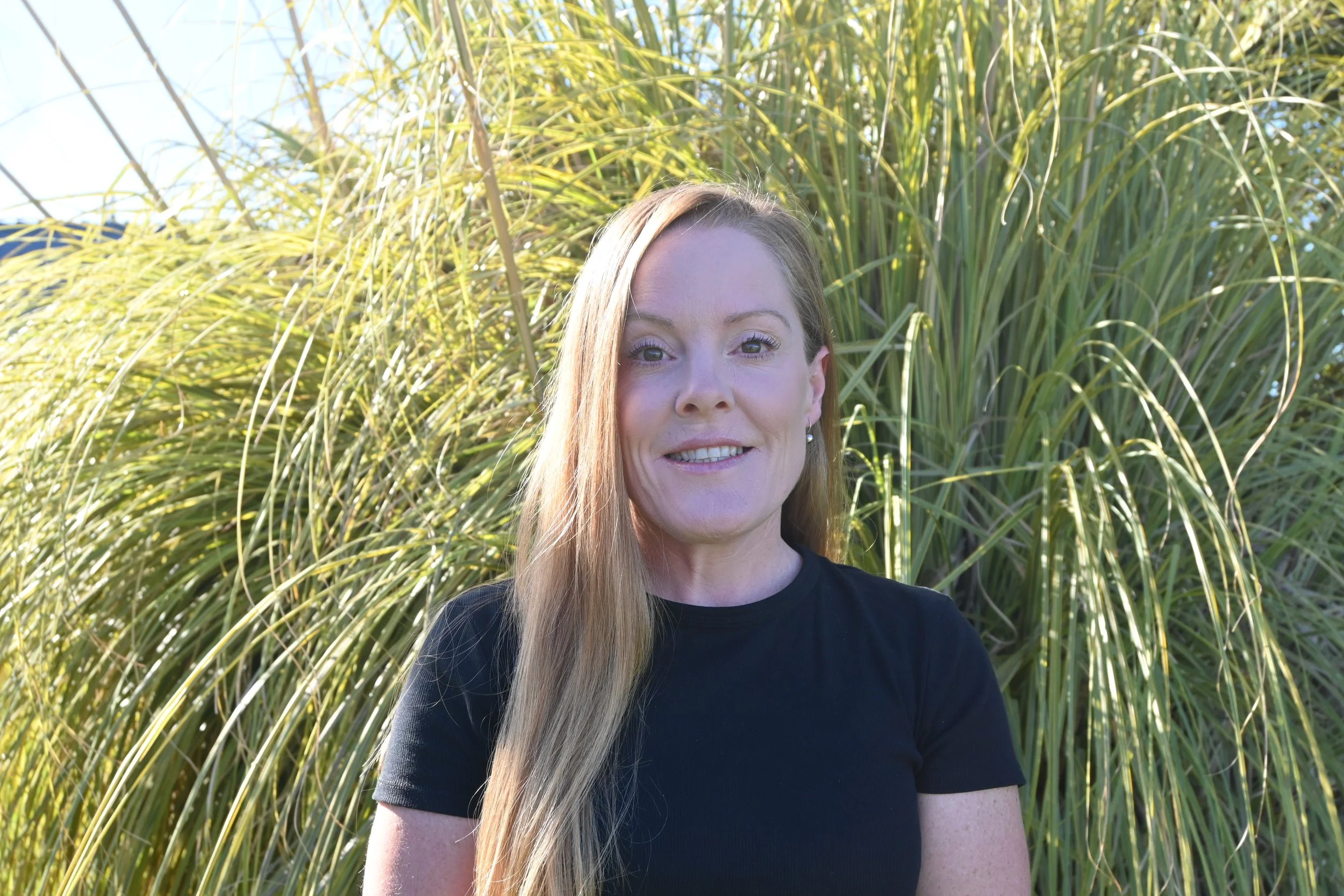 A woman with long blonde hair smiling outdoors in front of tall green grass.