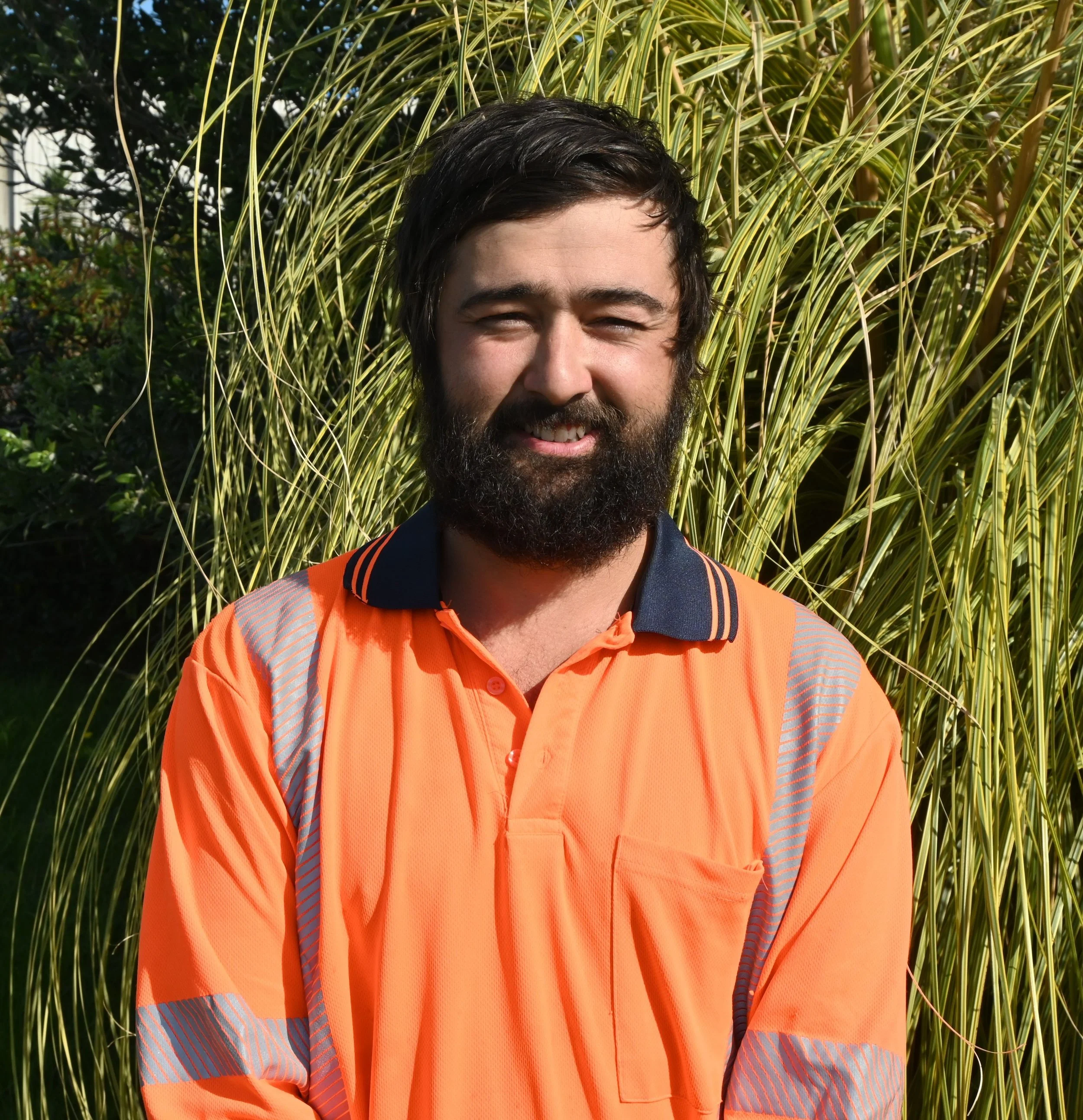 A man with dark hair and a beard, smiling, wearing an orange high-visibility work shirt, standing outdoors in front of tall, yellowish-green decorative grasses.