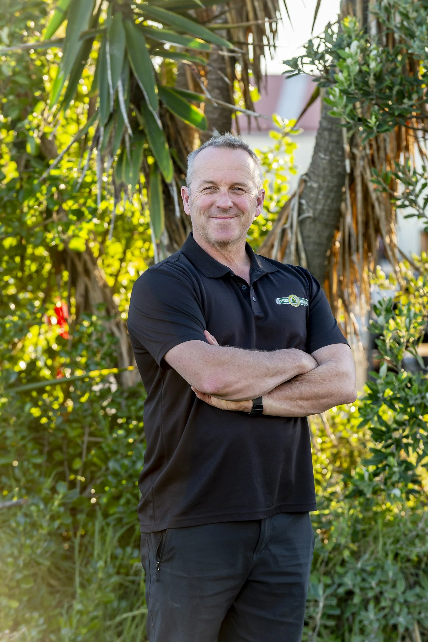 A man wearing a black polo shirt with a Tree Truck logo standing outdoors in front of green plants