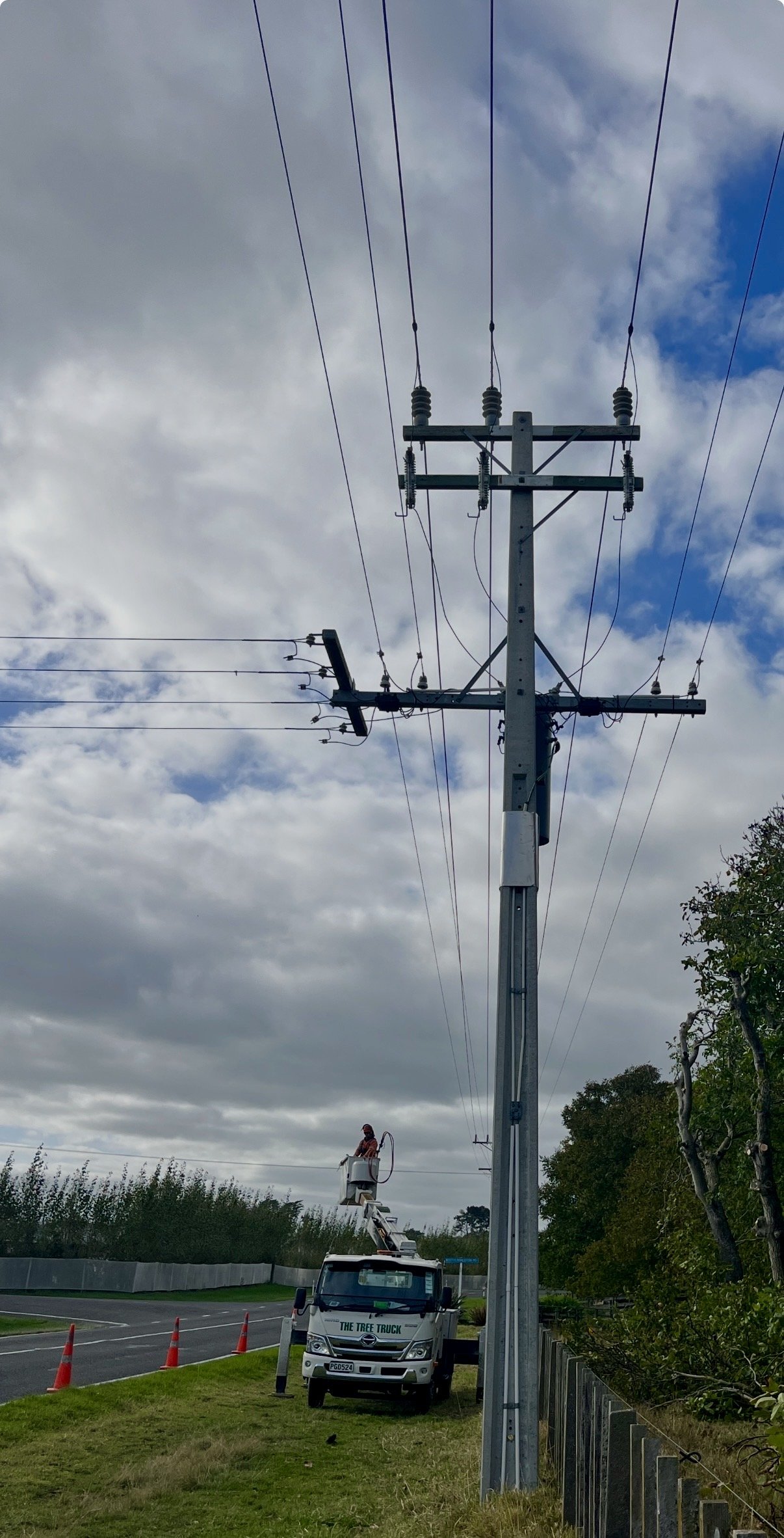 Utility worker on bucket lift repairing or maintaining a tall electrical power pole on the side of a road with orange traffic cones. Overcast sky with clouds.