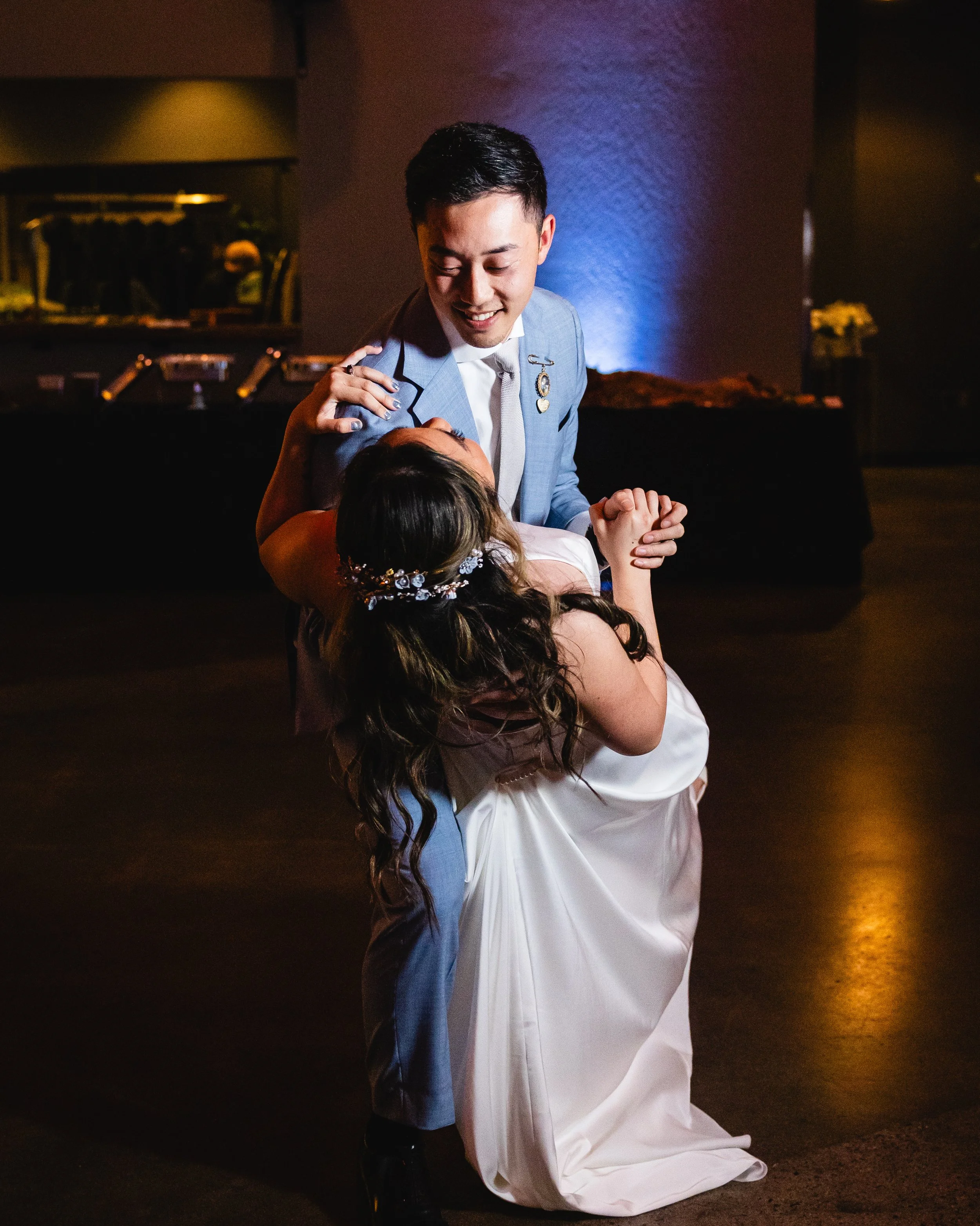 A man and woman dance together at a formal event, with the man holding the woman who is wearing a white gown and a floral headpiece.