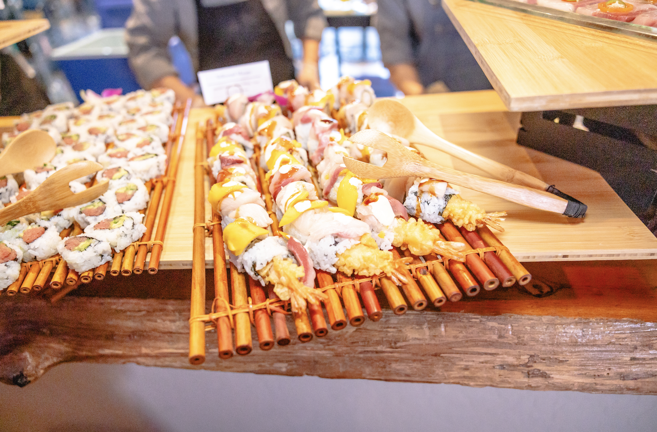 Assorted sushi rolls on bamboo mats at a sushi buffet with serving tongs.
