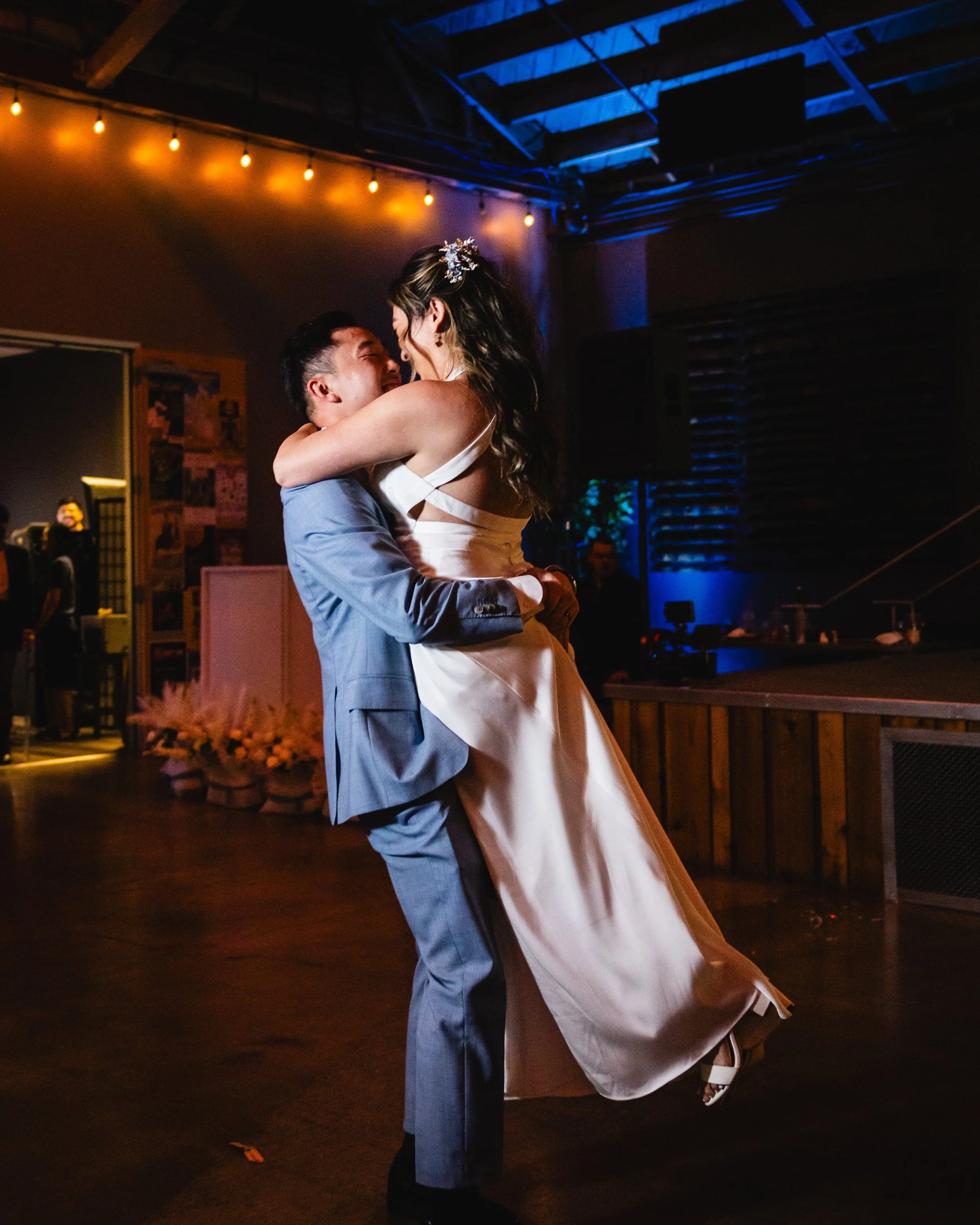 A couple dancing at a wedding reception, with the man lifting the woman in a white dress, surrounded by dim, warm lighting.