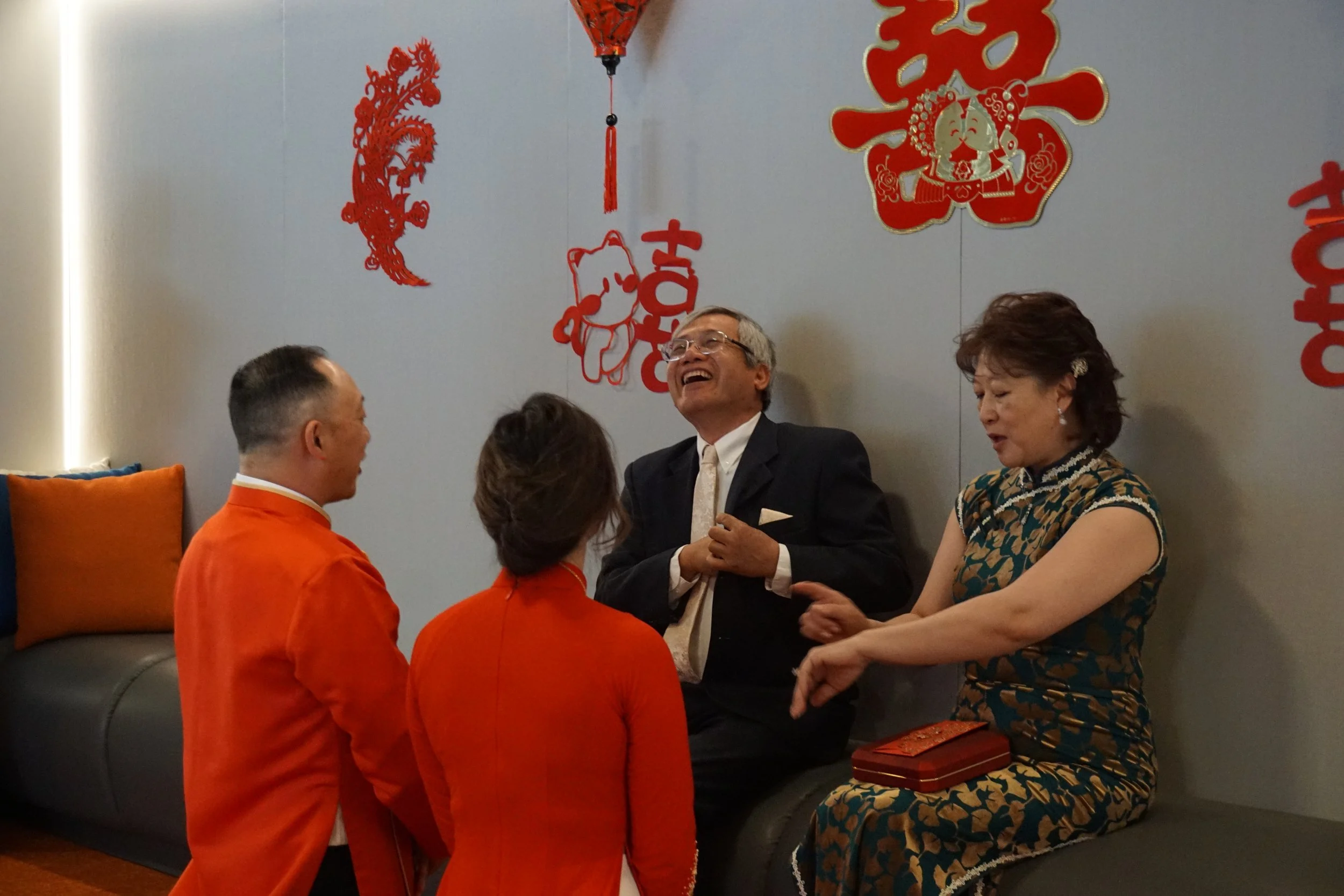 Four people, two women and two men, are having a joyful conversation in a room decorated with Chinese New Year symbols, such as red lanterns and wall hangings, with a gray wall background and cushions on a sofa.