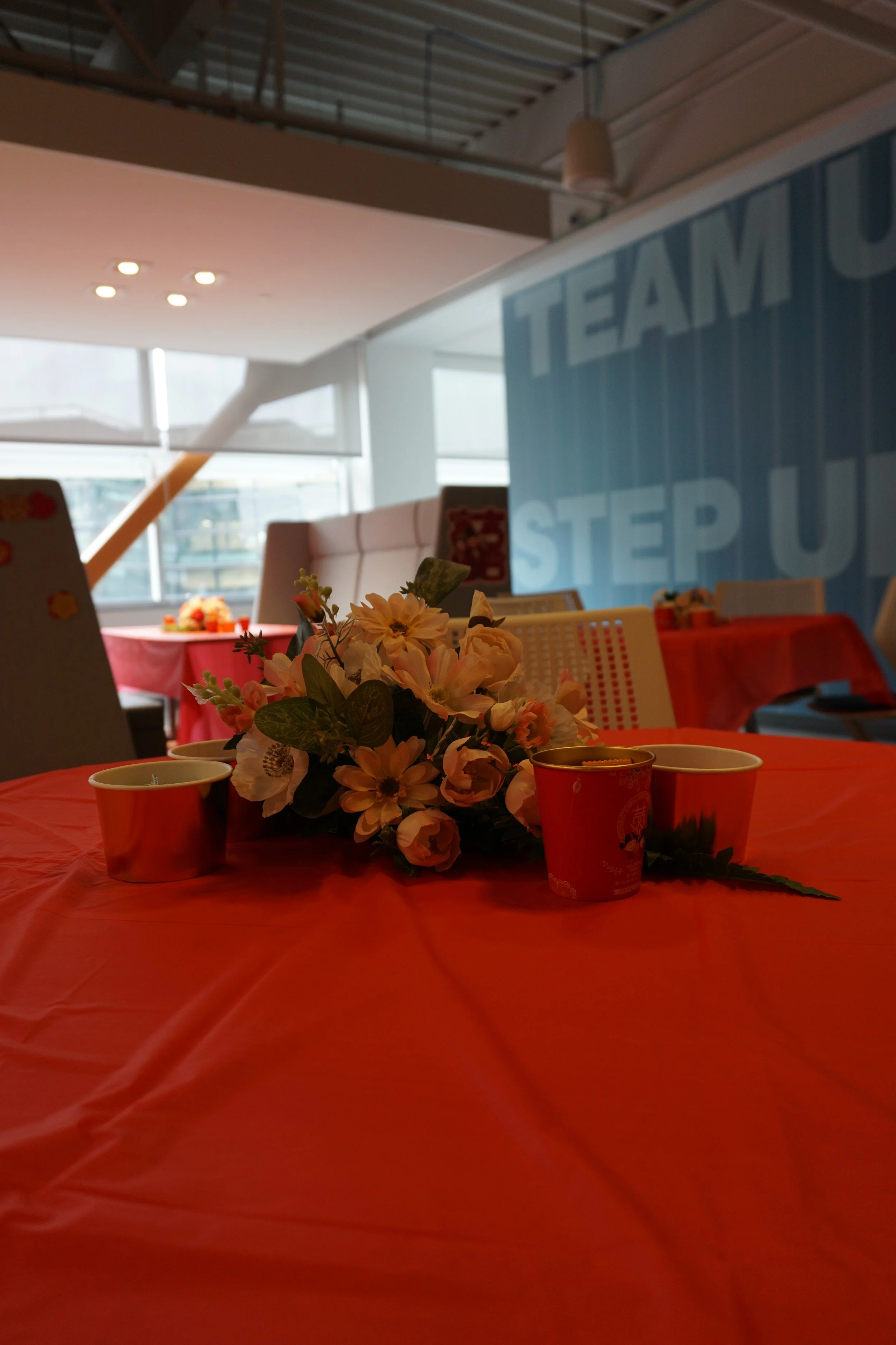 Table decorated with a pink tablecloth, a floral centerpiece, three red cups, and a teal banner with partially visible white text reading 'TEAM UP STEP U' in a room with large windows.