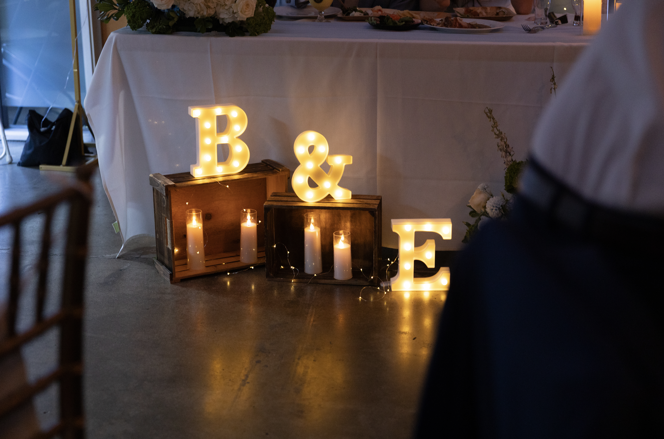 Decorative light-up letters 'B', '&', and 'E' on wooden crates with white candles and string lights at a wedding reception.