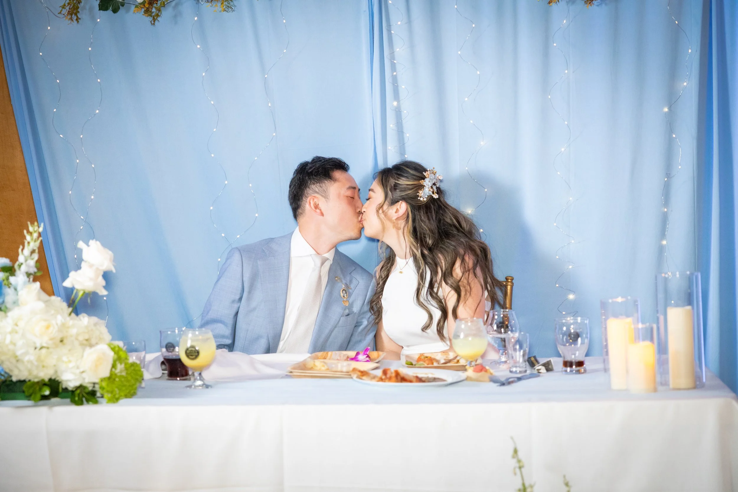 A bride and groom sharing a kiss at their wedding reception, sitting at a decorated table with flowers, drinks, and candles.