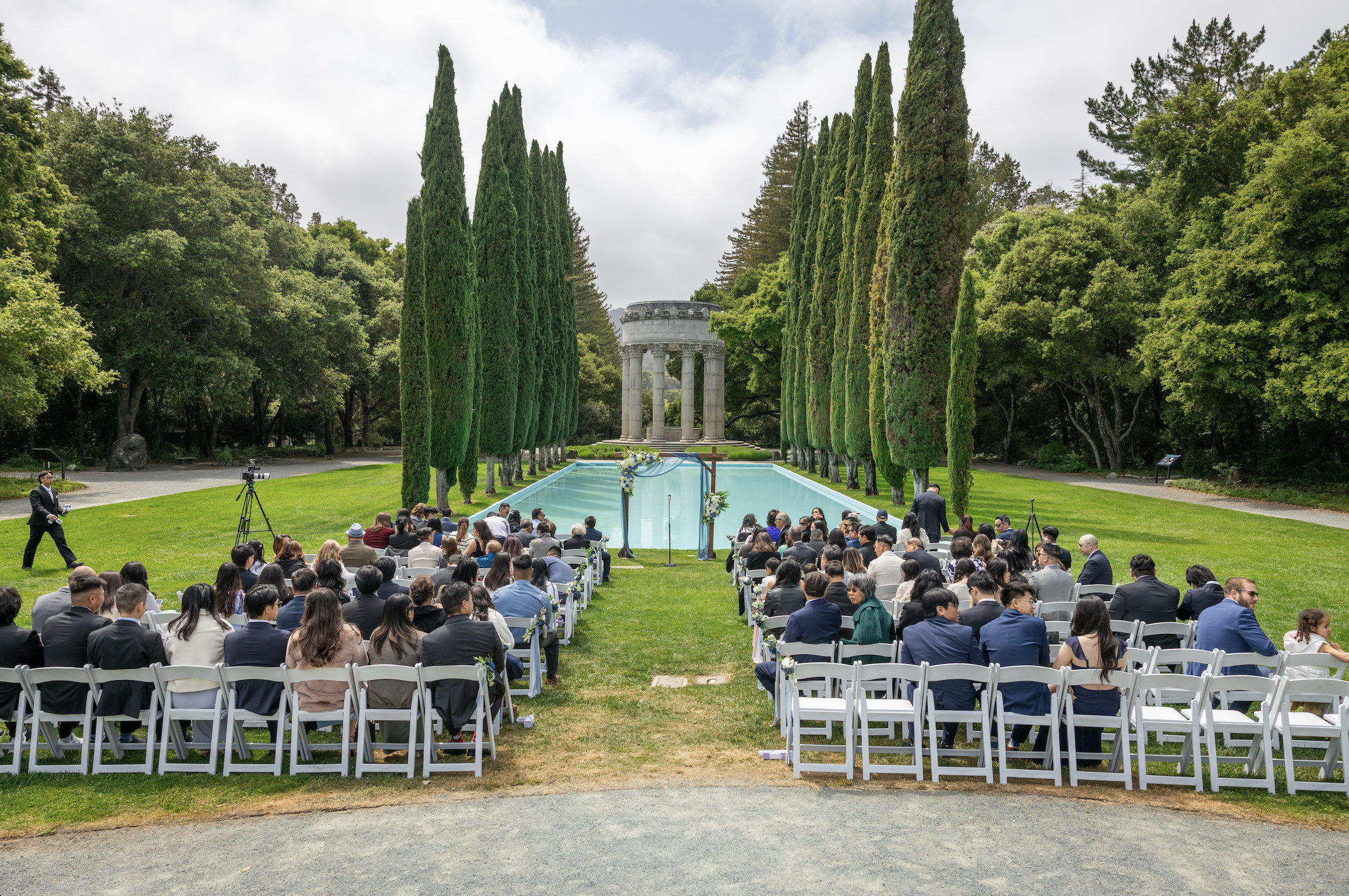 Outdoor wedding ceremony with guests seated on white chairs facing a decorated arch near a rectangular pool, surrounded by tall cypress trees and lush greenery, with a classical monument in the background.