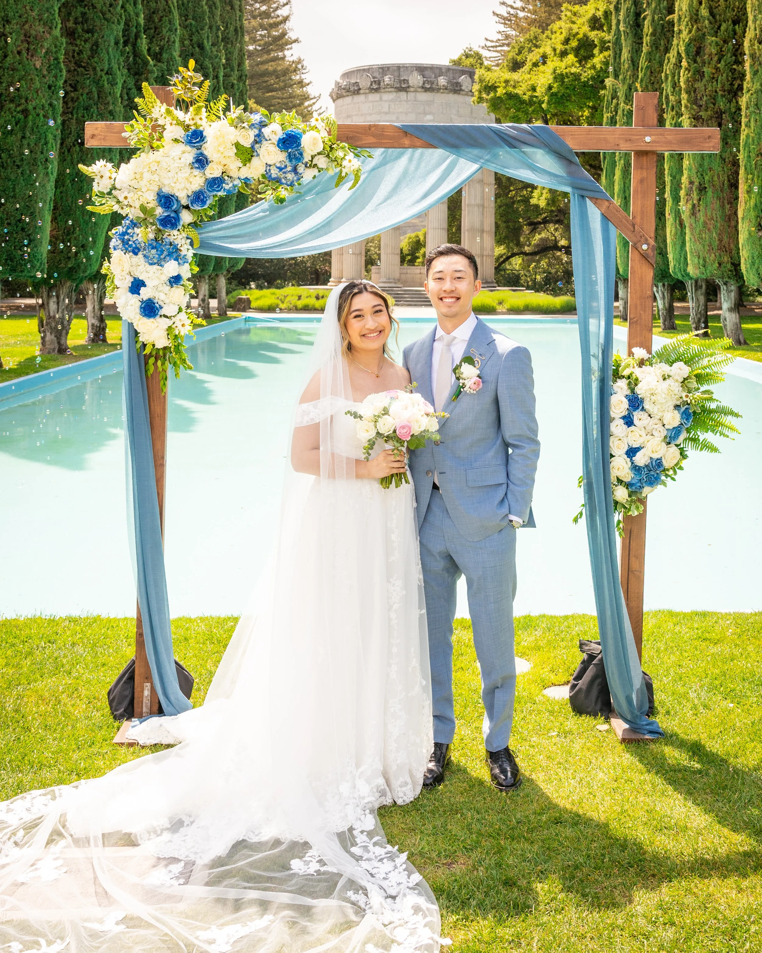 Happy bride and groom standing under wedding arch with blue and white flowers, outdoors near a pond and historic columns.