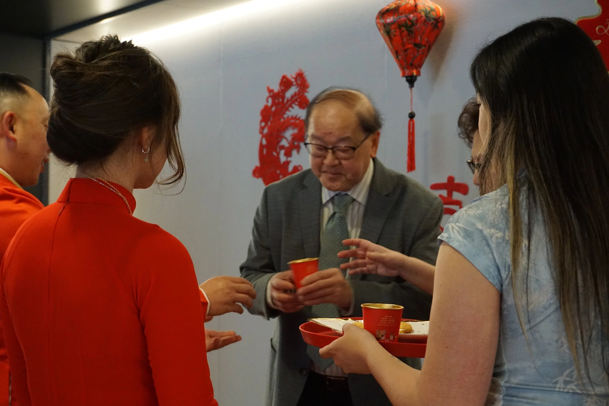 A group of people in traditional Chinese attire and formal wear are engaging in a tea or drink exchange during a celebration, with decorations including a red lantern and Chinese characters in the background.