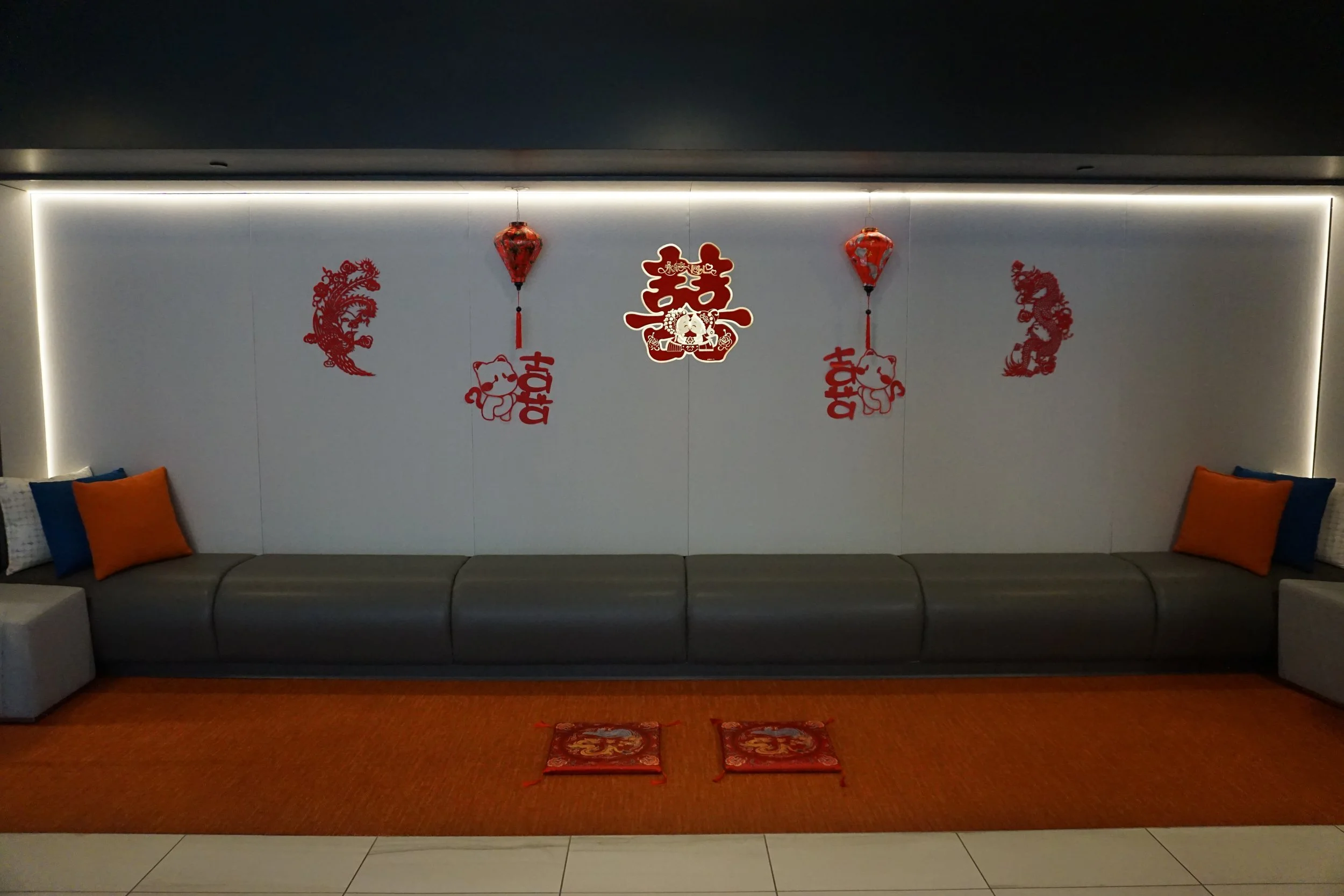 Decorations for Chinese New Year or celebration with red Chinese lanterns, red paper cutouts of dragons, the word 'Happy' and a double happiness symbol on a white wall above a long gray sofa with orange and blue pillows.