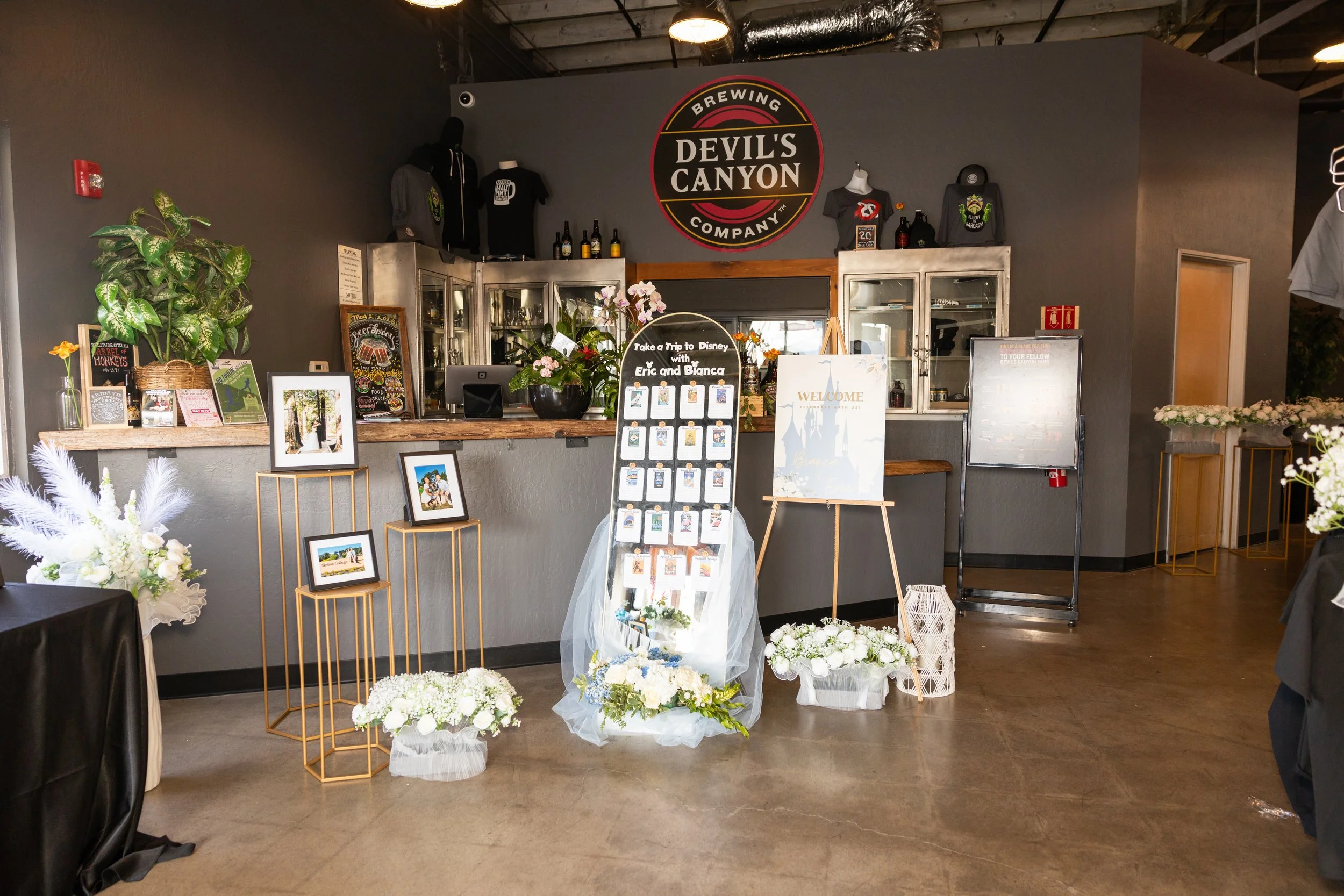 Interior of a brewery with a gray wall and metal counter. Signs read "Devil's Canyon Brewing Company" and "Take a Trip to Disney with Eric and Bianca." Decor includes framed photos, flower arrangements, and a large plant.