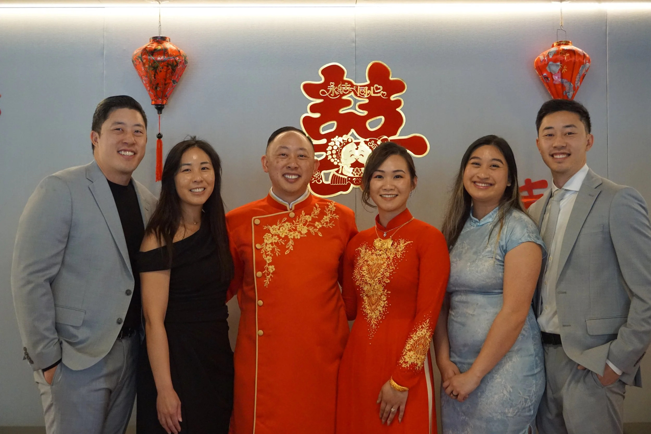 Group of seven people posing for a photo during a celebration with traditional Chinese decorations, including red lanterns and a large red and gold cutout with Chinese characters in the background.
