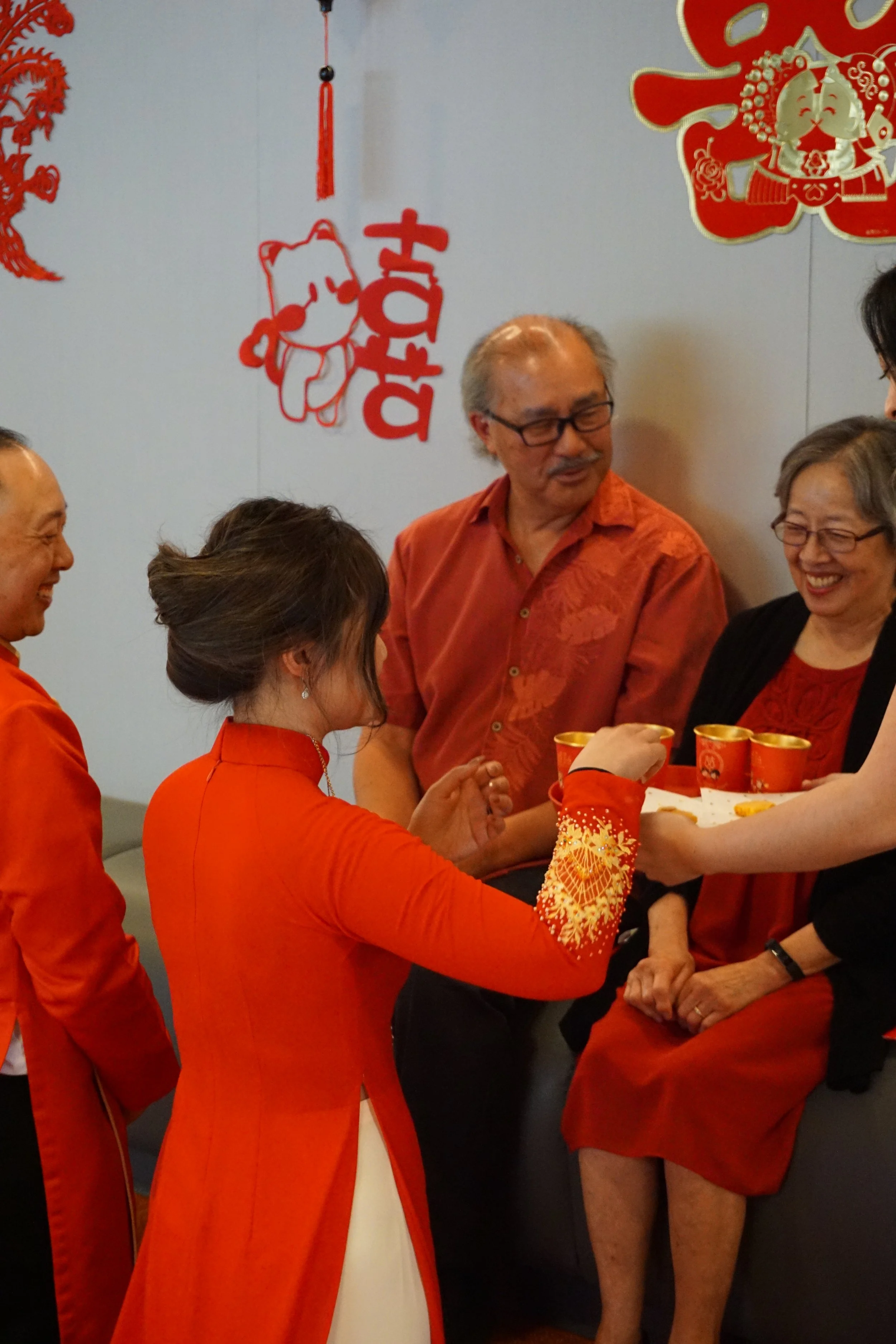 A group of people celebrating Lunar New Year, dressed in red and black clothing, with red and gold decorations and a 'Happy New Year' sign on the wall. They are sharing drinks and smiling.