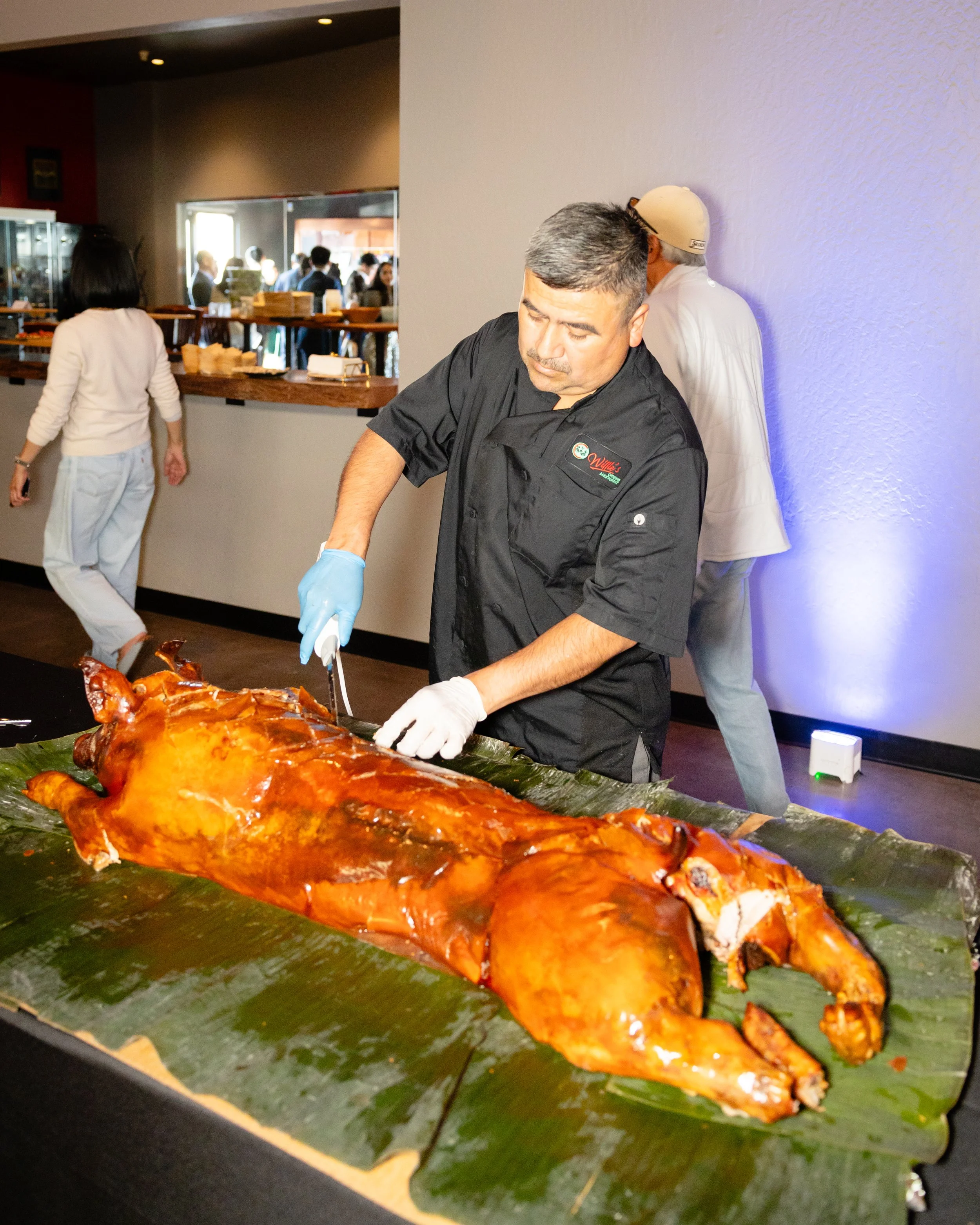 A chef wearing a black uniform and blue gloves carves roasted whole pig on a table lined with banana leaves at a buffet or event.