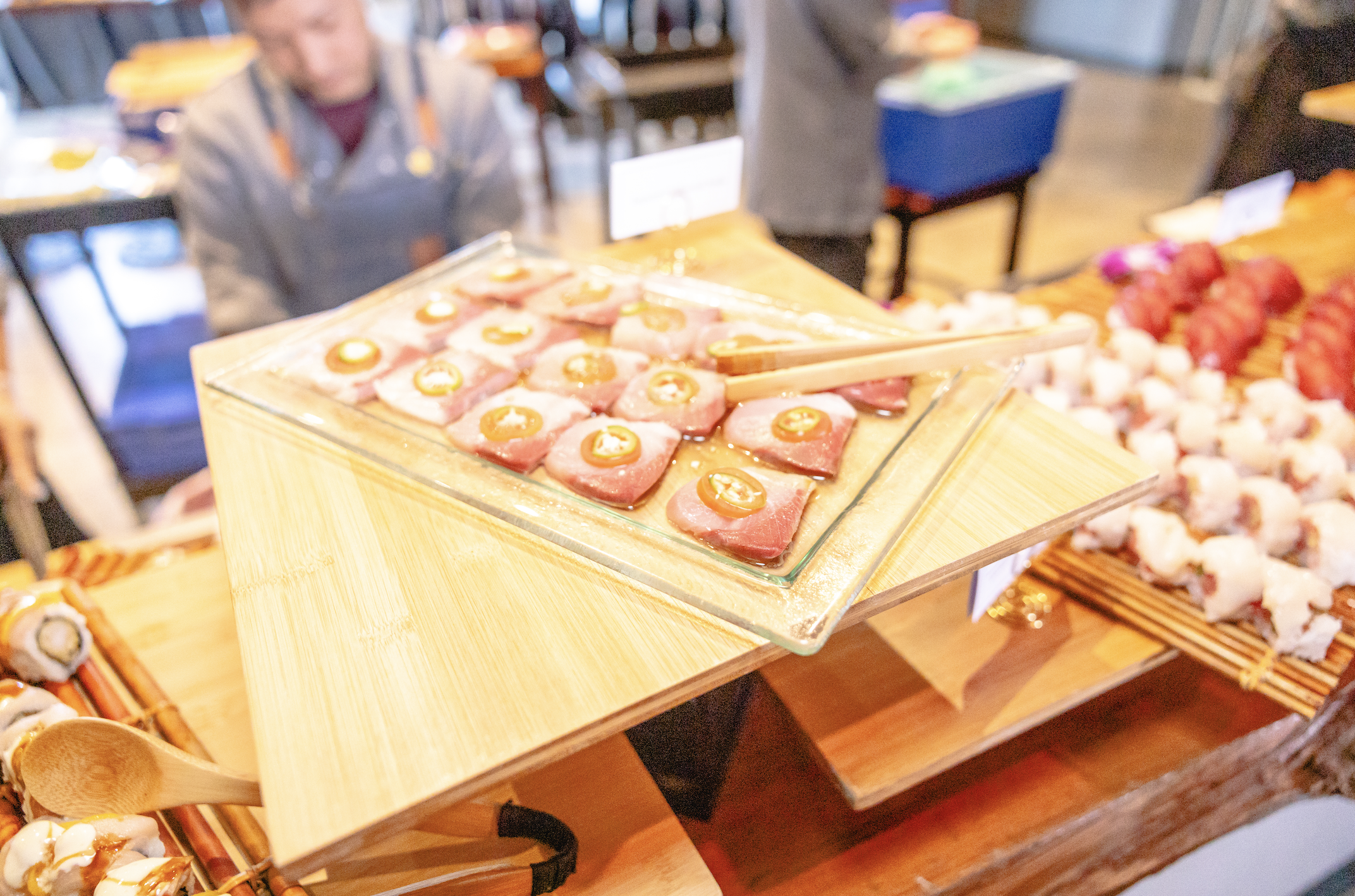 Close-up view of a glass tray with pieces of sushi topped with slices of yellow chili pepper, set on a wooden display table at a buffet or restaurant.