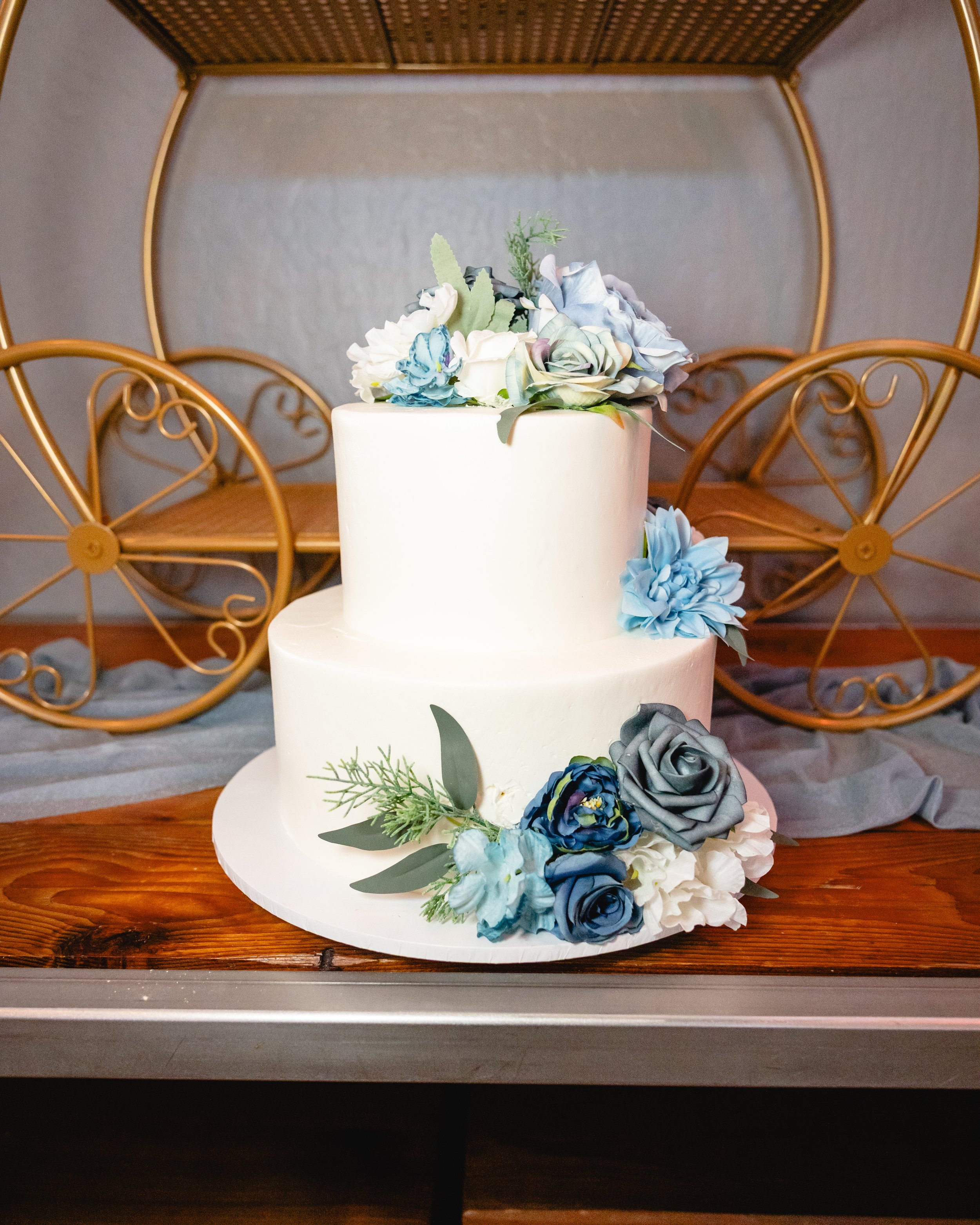 Two-tier white wedding cake decorated with blue, gray, and green flowers, placed on a wooden table with a vintage golden carriage in the background.