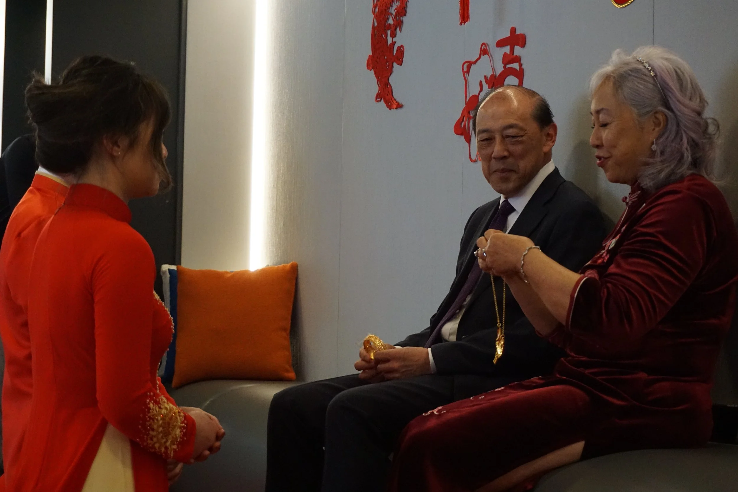 A woman in traditional Vietnamese dress and two older adults, a man and a woman, sitting on a couch, engaged in a conversation indoors, with red decorations on the wall.