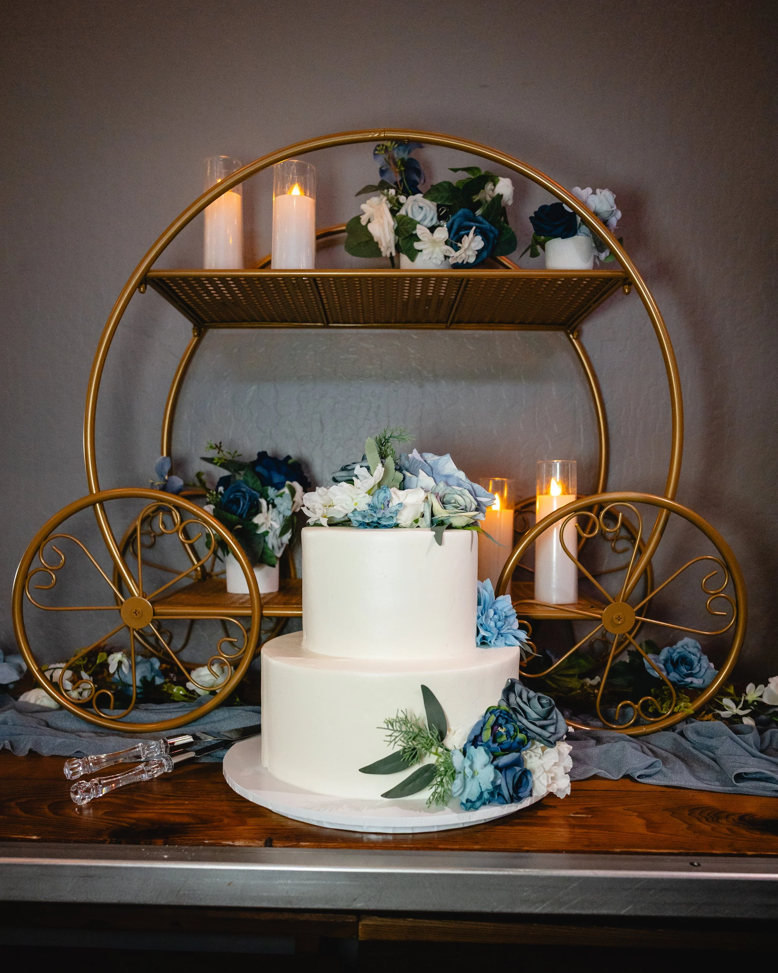 A two-tier white wedding cake decorated with blue and white flowers, placed on a wooden table. In the background, there is a decorative gold cart with candles and floral arrangements.