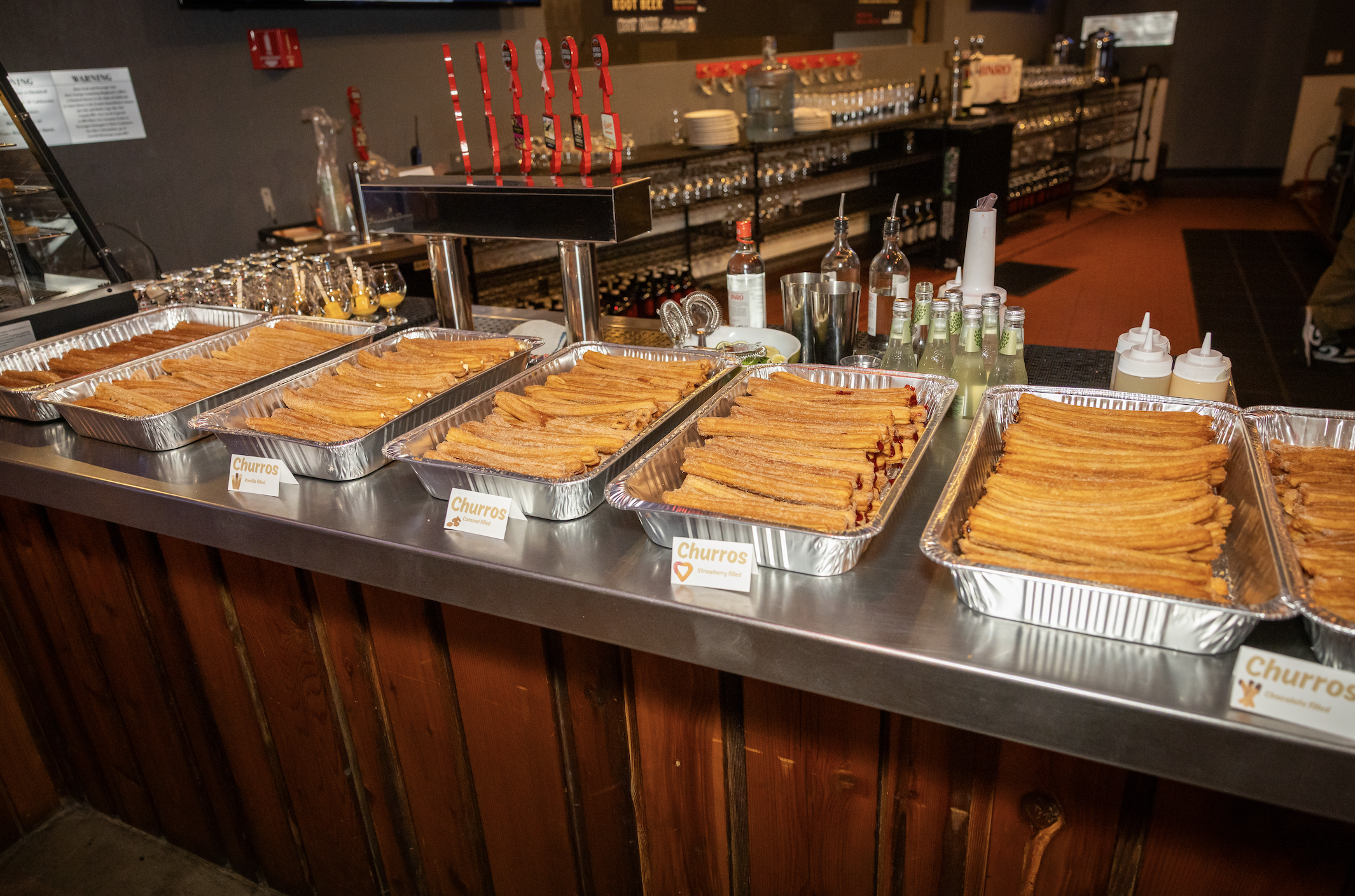 Several trays of churros on a counter at a restaurant or café, with bottles of syrup and condiments in the background.
