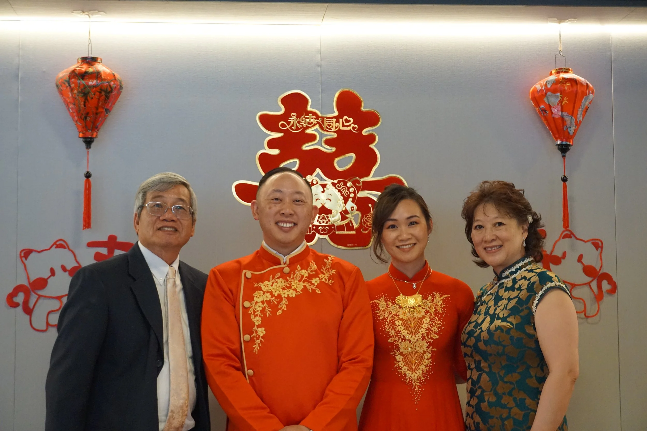 Four people in traditional Chinese attire and formal suit posing in front of a wall decorated for Chinese New Year, with red lanterns and Chinese characters in the background.