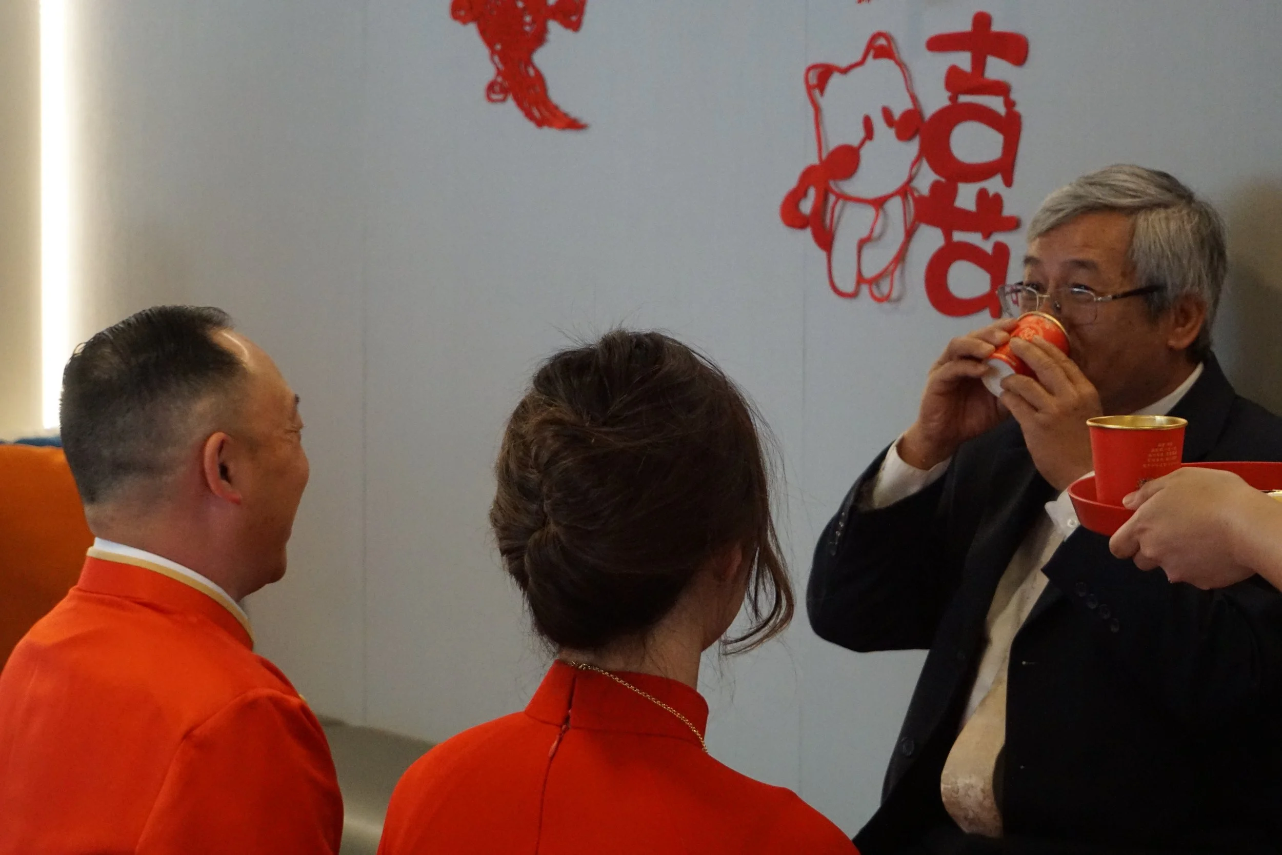 Three people, two men and a woman, dressed in traditional red and orange clothing, are participating in a tea ceremony. The man on the right is drinking from a small red cup, while the other man and woman are watching and smiling. The background show